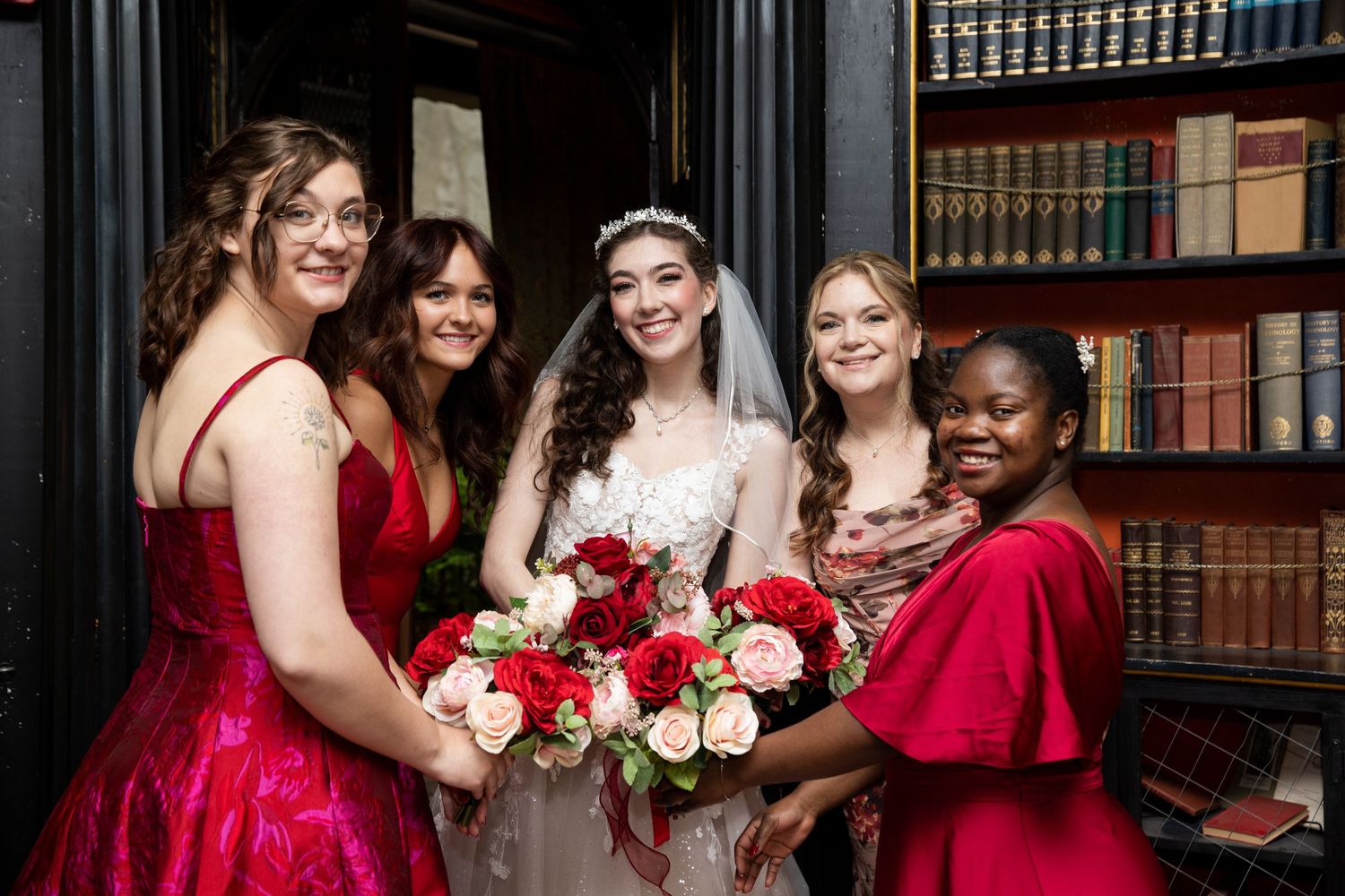 A bridal party poses in a library wearing burgundy dresses and holding red and pink rose bouquets.