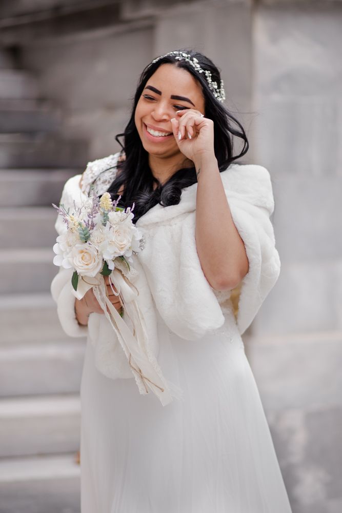 Joyful bride wearing white winter dress and delicate hair accessories holds pastel bouquet while wiping happy tears.