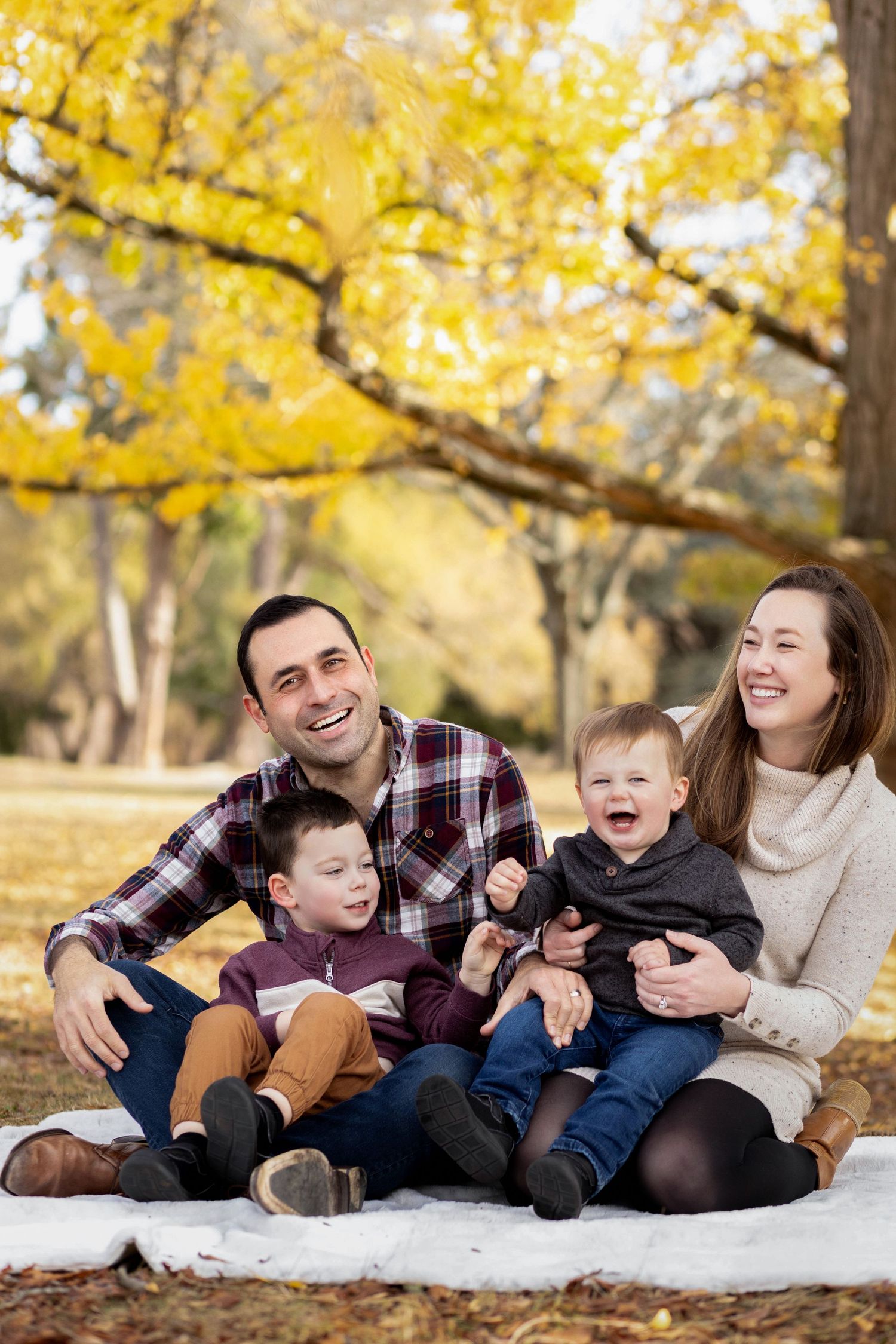 A happy family sits together on a blanket outdoors during autumn with golden yellow foliage in the background.