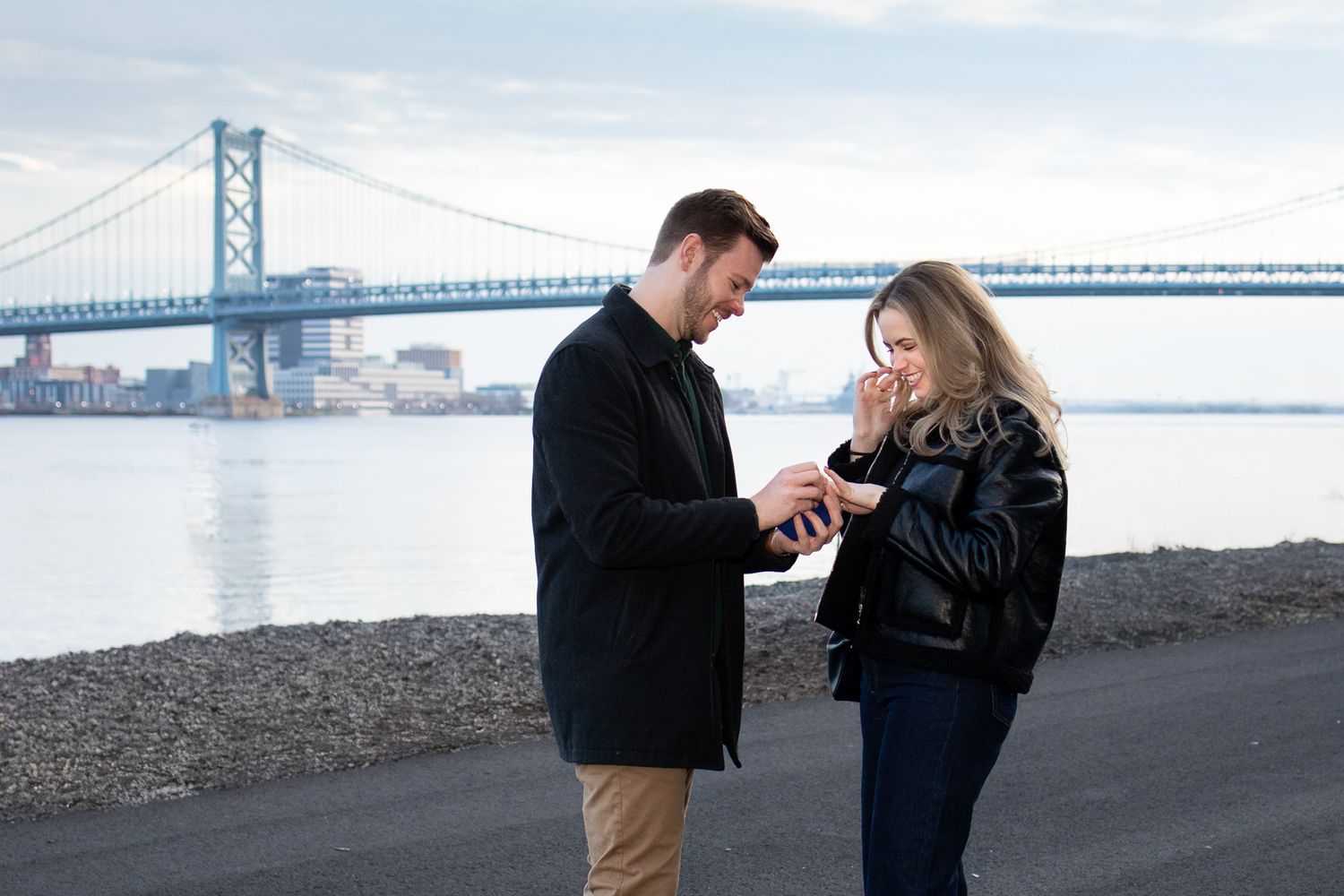 A couple shares a romantic moment near a bridge with the San Francisco Bay Bridge visible in the background.