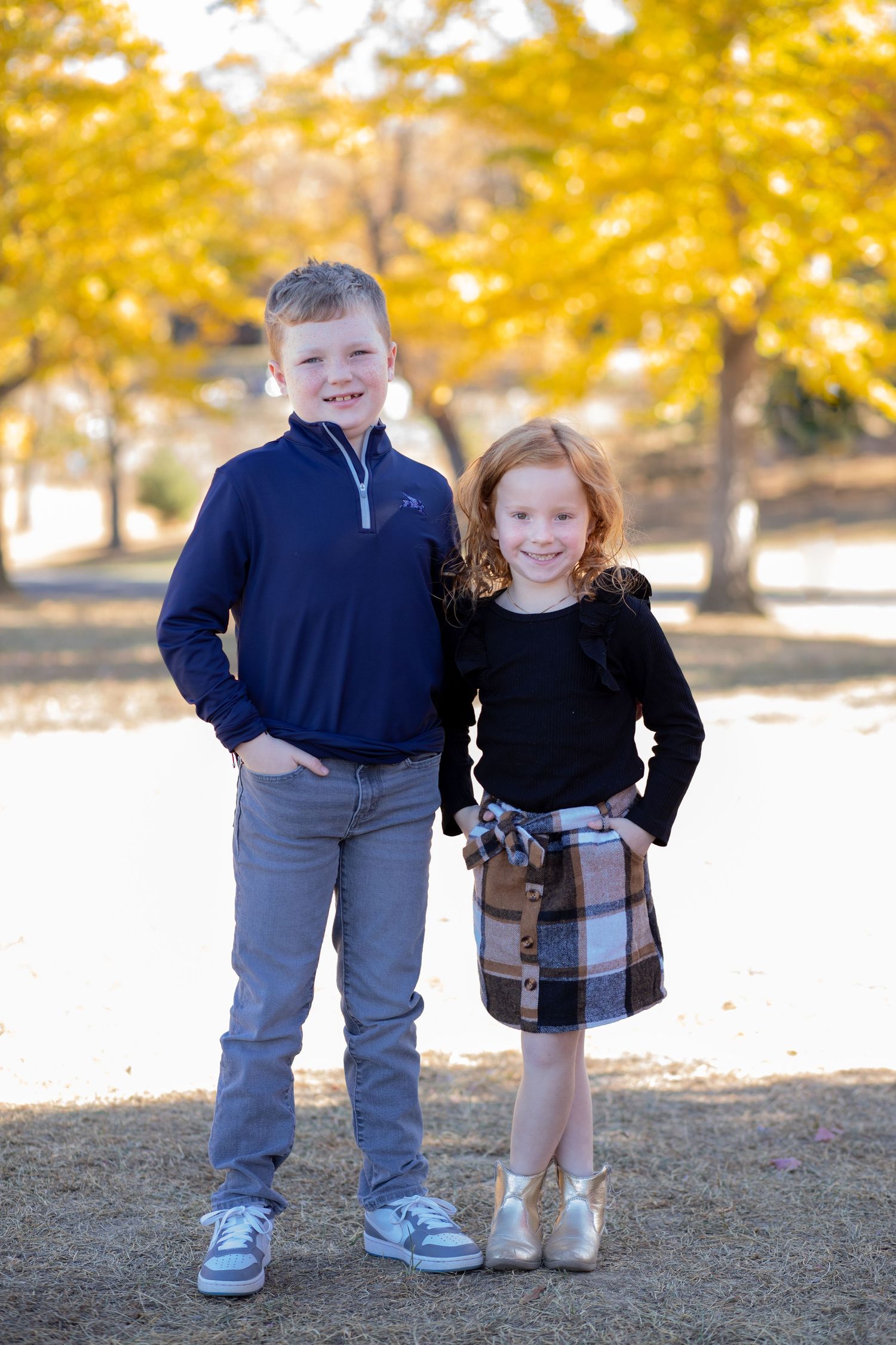 Two children stand outdoors in fall, wearing dressy casual clothes with golden autumn trees in the background.