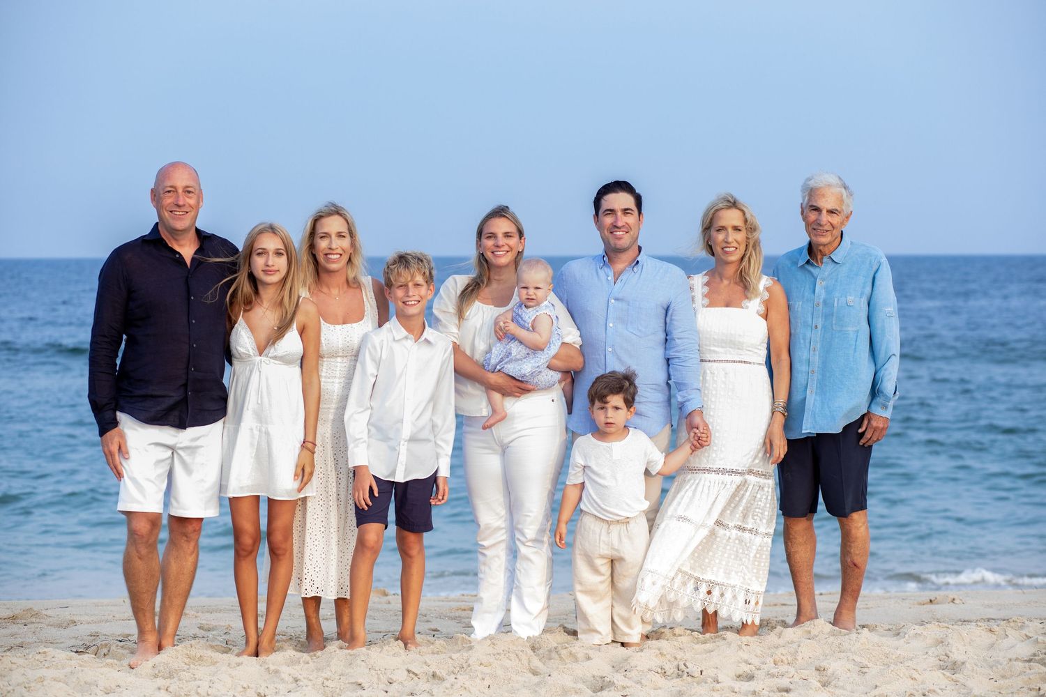 A group in coordinated white and blue outfits poses together on a sunny beach with ocean waves in the background.