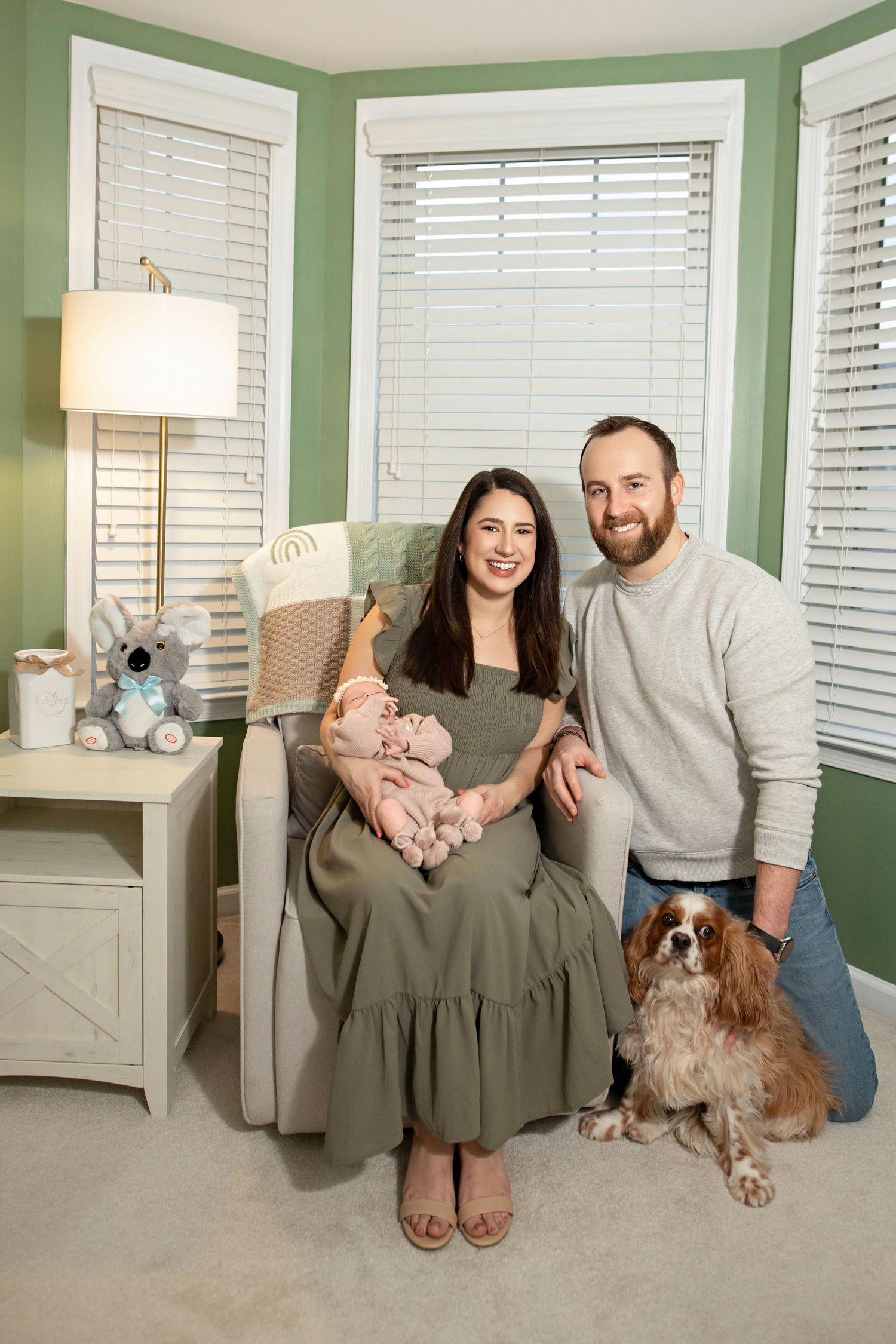 A cozy nursery corner with sage green walls featuring a comfortable chair, pet dog, and white window blinds.