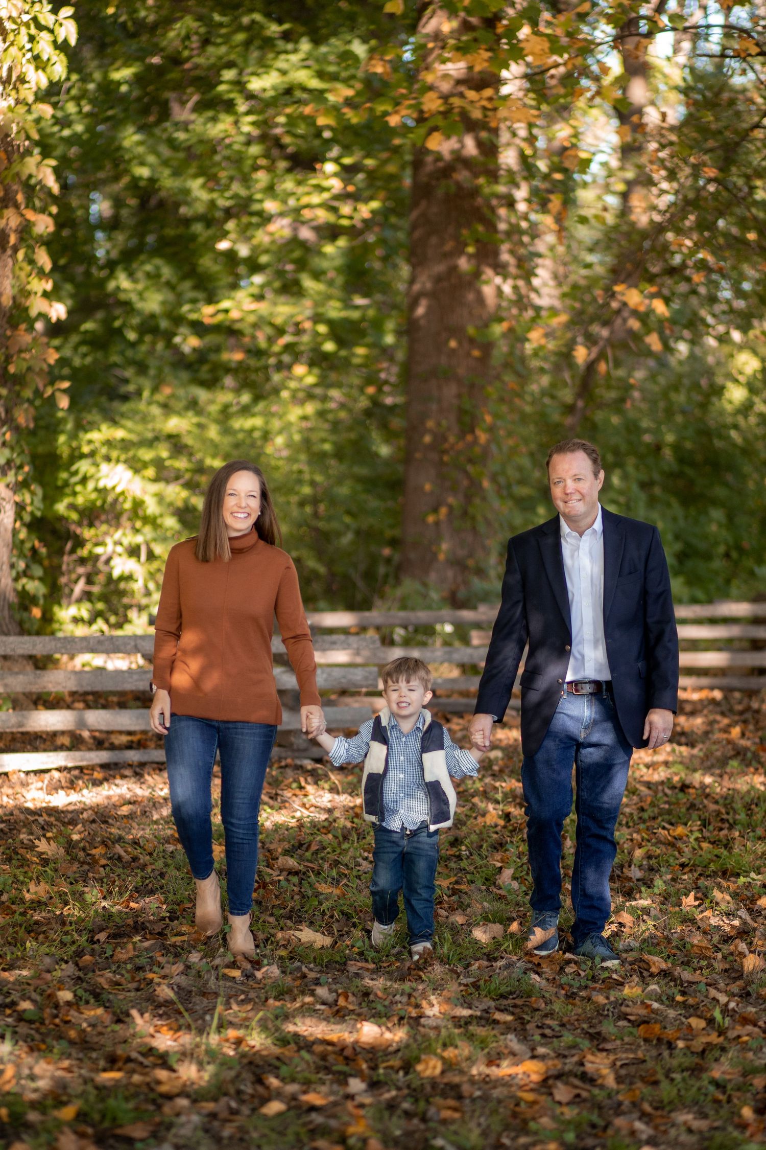 A family walks together on a leaf-covered path through a wooded area on a sunny autumn day.