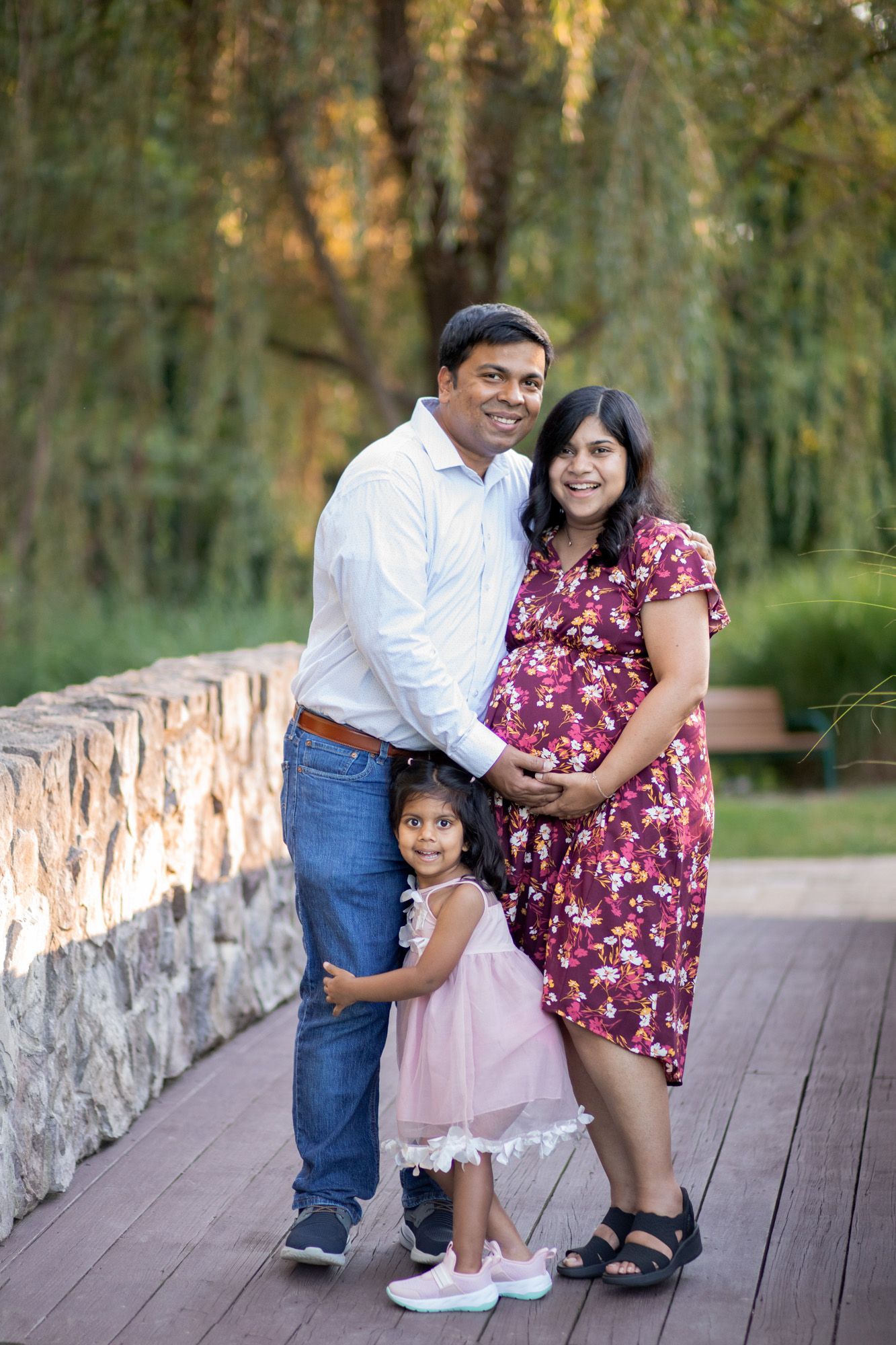 A happy Indian family poses outdoors, with a pregnant mother in a floral dress, father, and young daughter on a wooden deck.