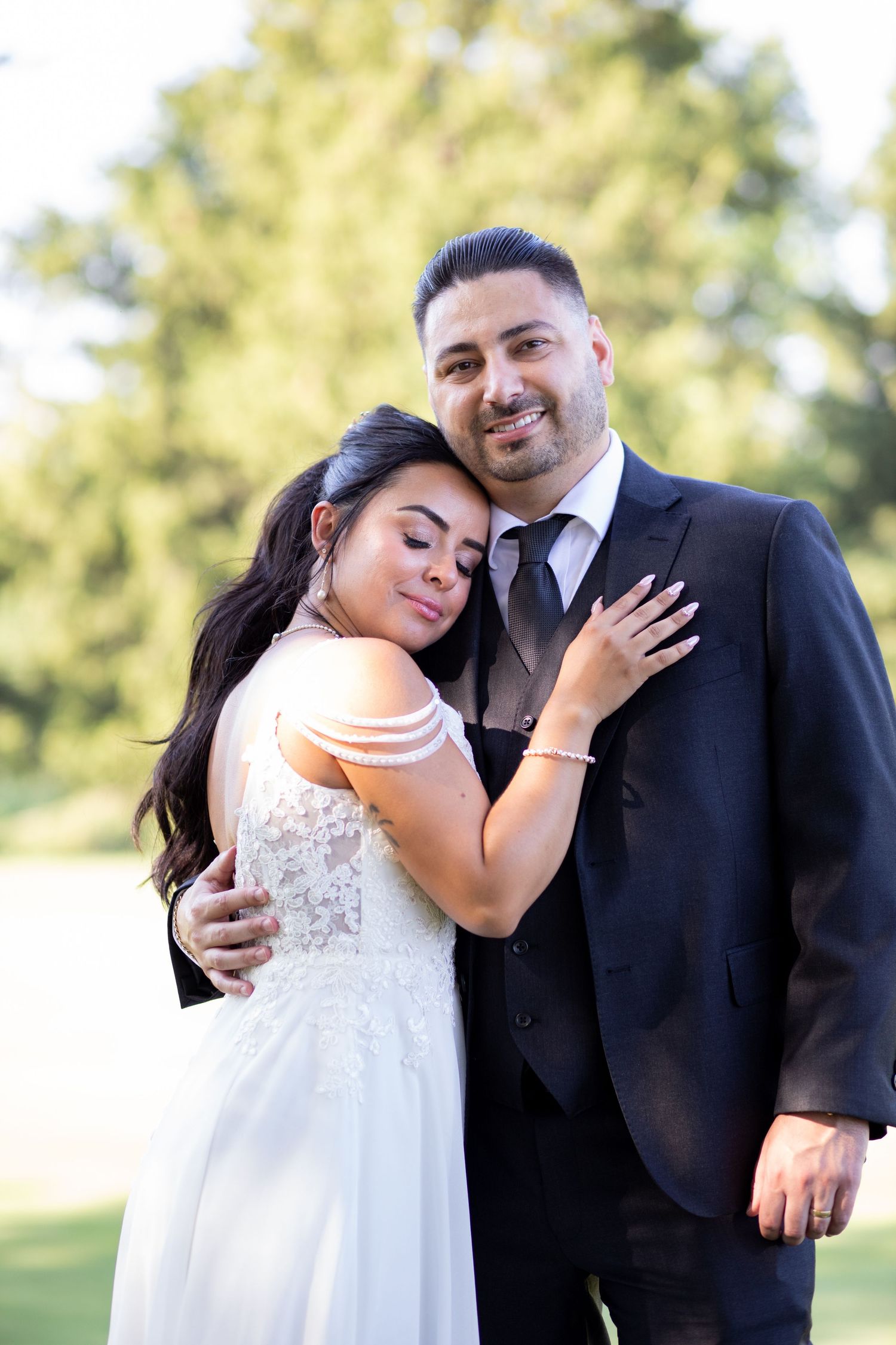 A couple embraces outdoors in formal wedding attire against a soft blurred background of trees.