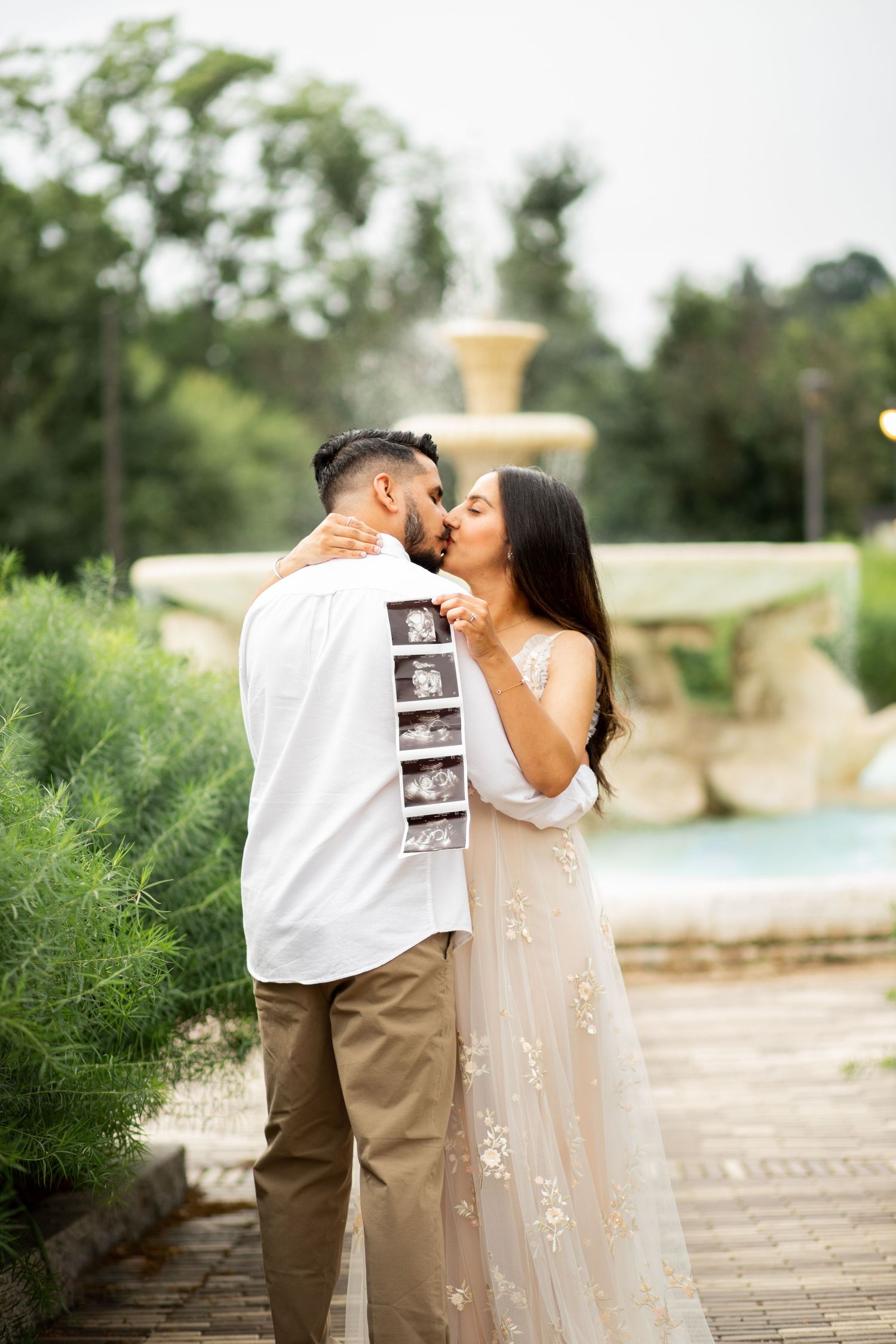 A couple embraces while sharing an ultrasound photo in an outdoor garden setting.