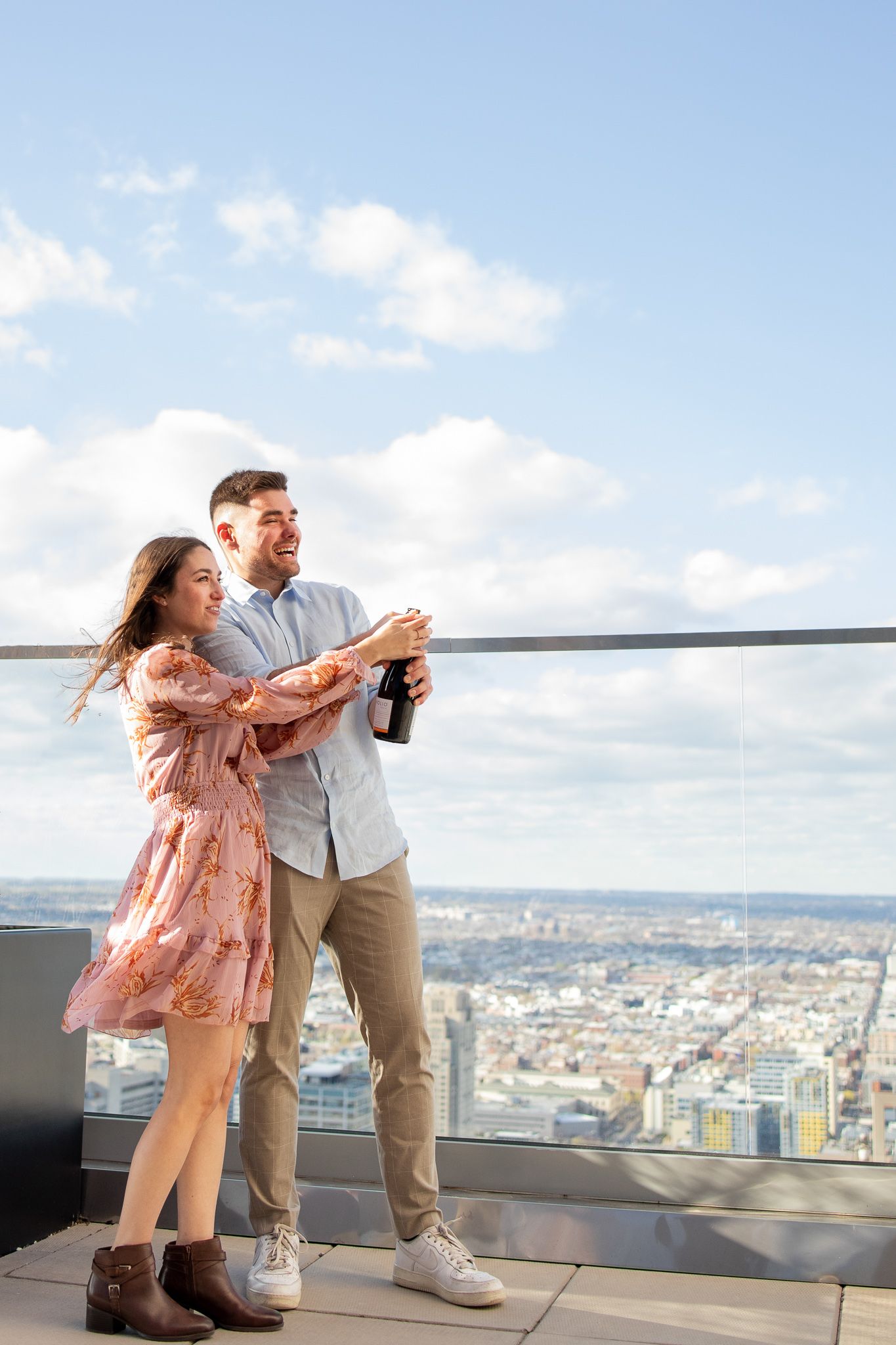 A couple enjoys a playful moment on an observation deck overlooking a cityscape on a sunny day.