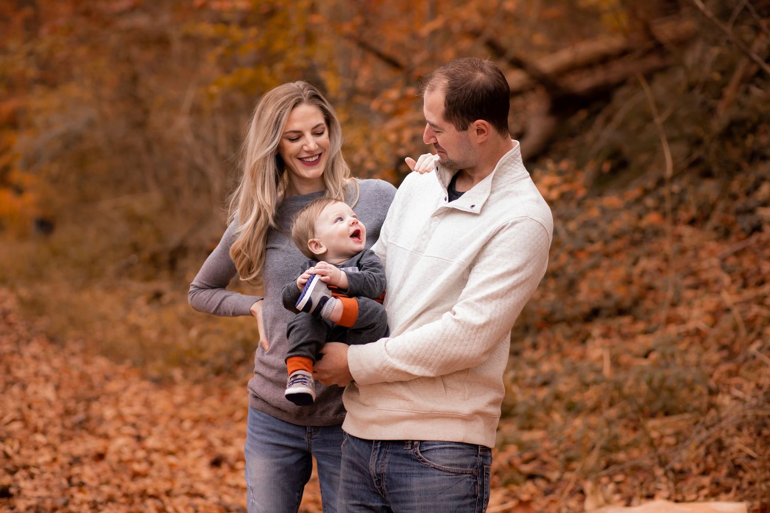 A couple and their baby share a happy moment together surrounded by autumn foliage in an outdoor forest setting.