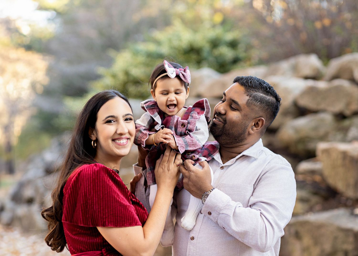 A family stands together outdoors in autumn, laughing and playing with their young child near stone formations.