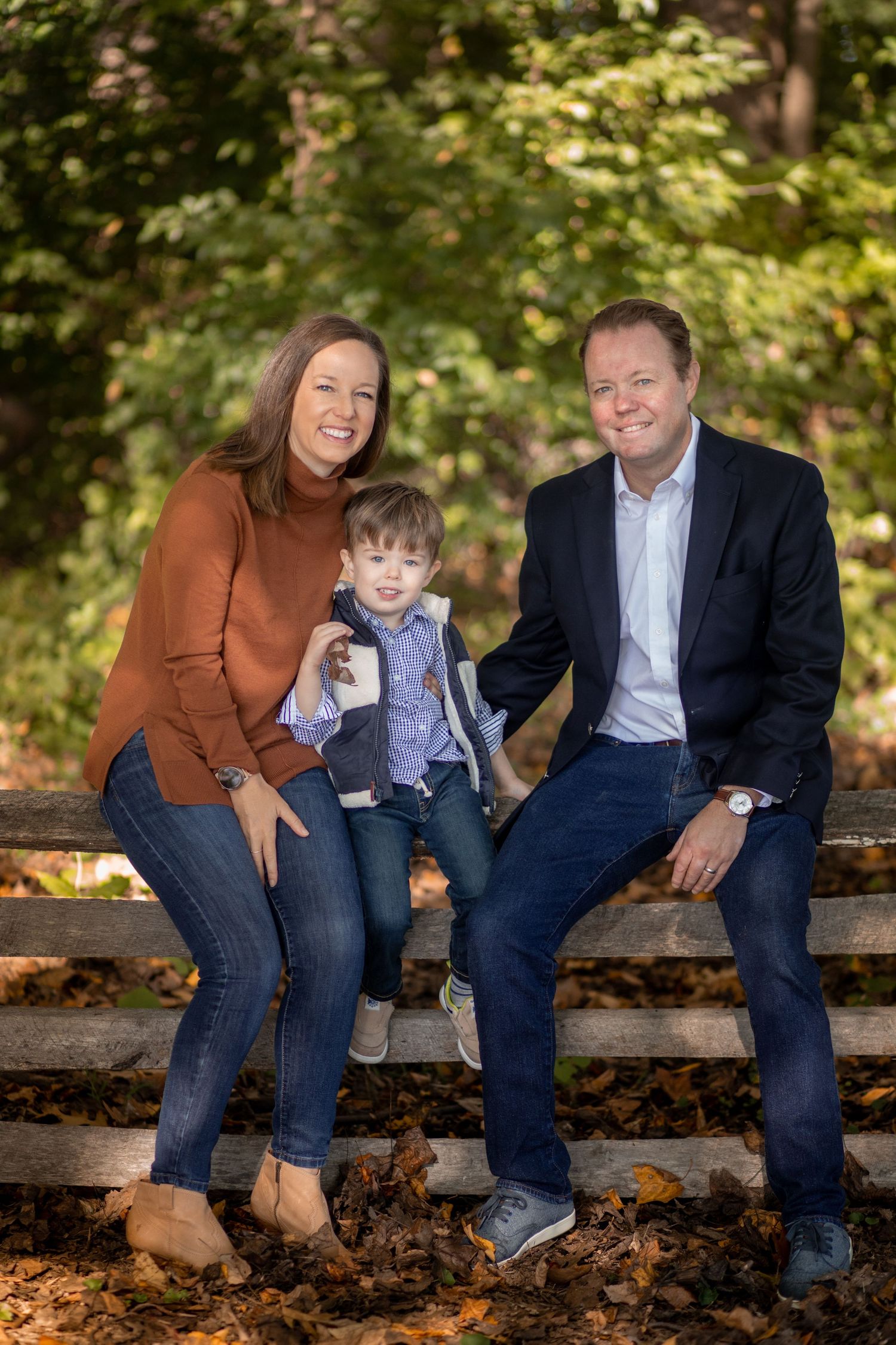 A family of three shares candid laughter while sitting on a rustic wooden fence surrounded by autumn foliage.