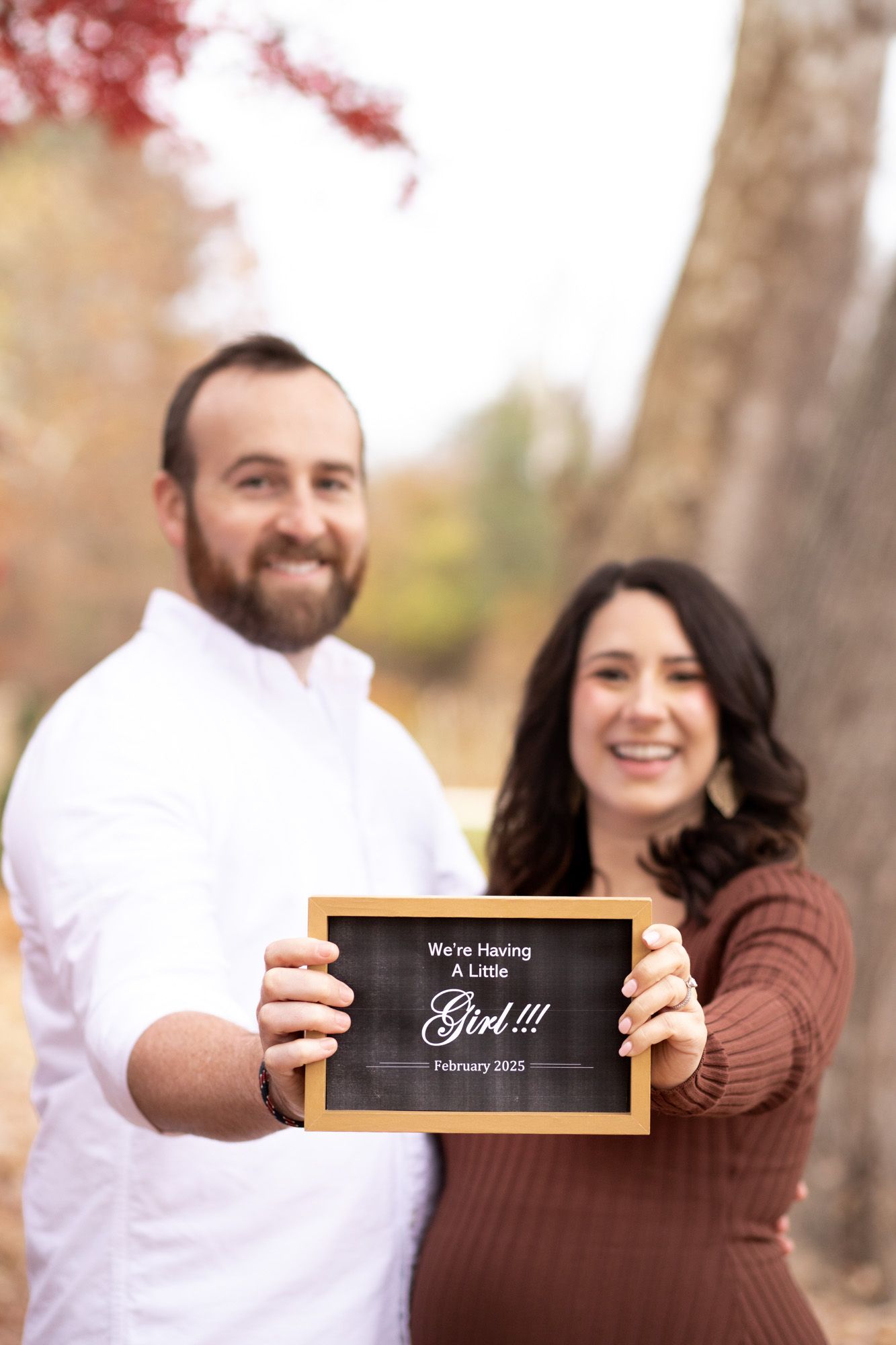 Happy couple holding a chalkboard sign announcing they are expecting a baby girl in February 2025.