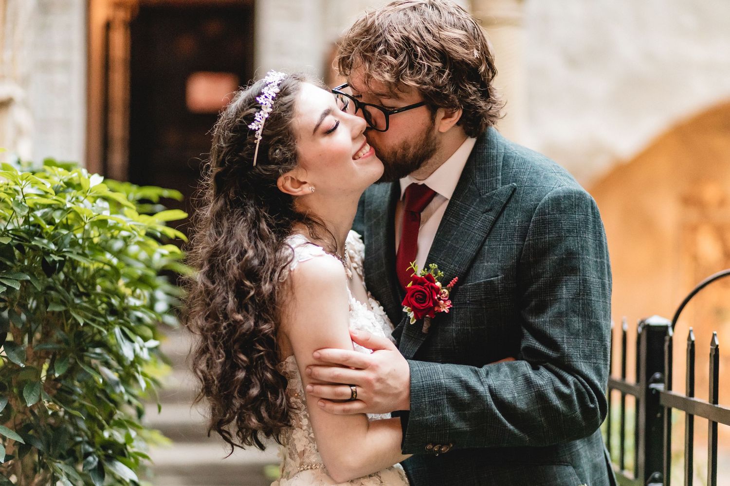 A romantic couple in wedding attire embrace intimately in a garden setting with greenery and sunlit stone walls.
