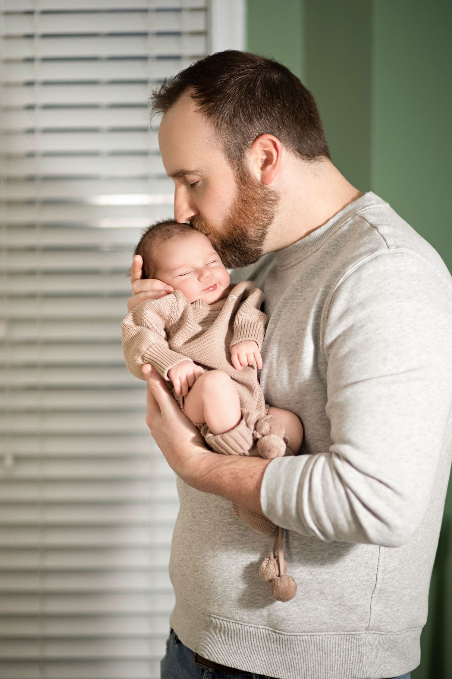 A tender moment as a parent in a gray sweater gently holds and kisses a sleeping newborn baby near a window.