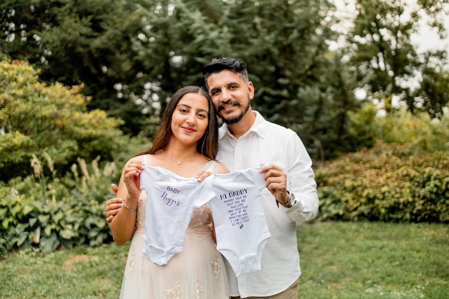 Two people in white outfits hold up matching embroidered handkerchiefs while standing in a sunny garden setting.