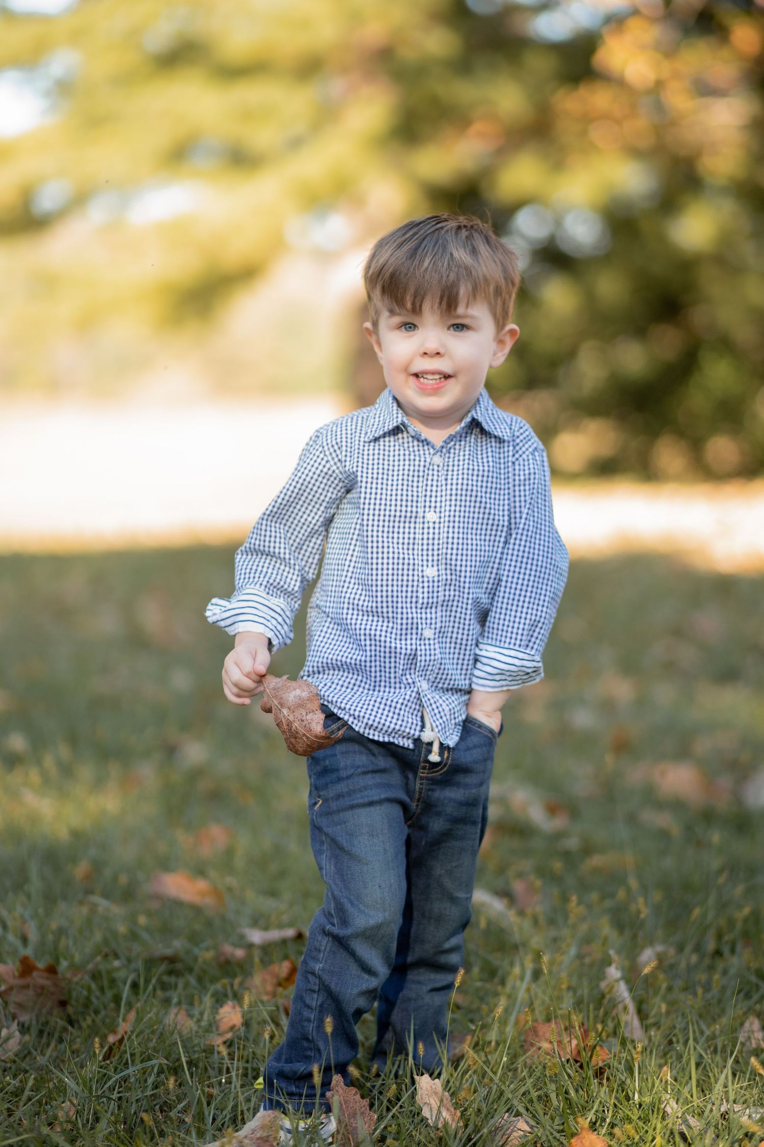 A child in a blue gingham shirt and jeans stands smiling on grass with autumn leaves and soft bokeh in the background.