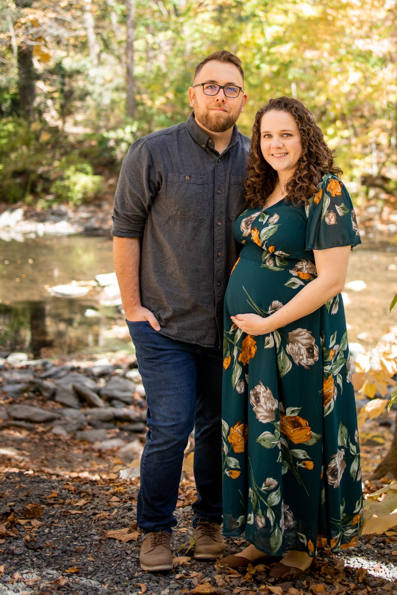 Pregnant couple posing outdoors near a creek surrounded by autumn foliage, woman in floral teal dress cradling baby bump.