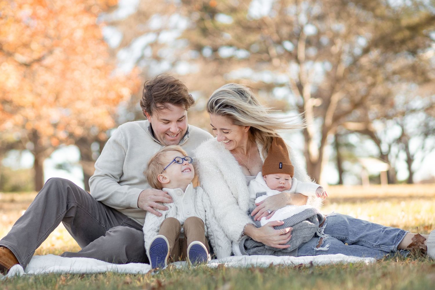 Family sitting together on blanket laughing and playing during autumn photo session in park.