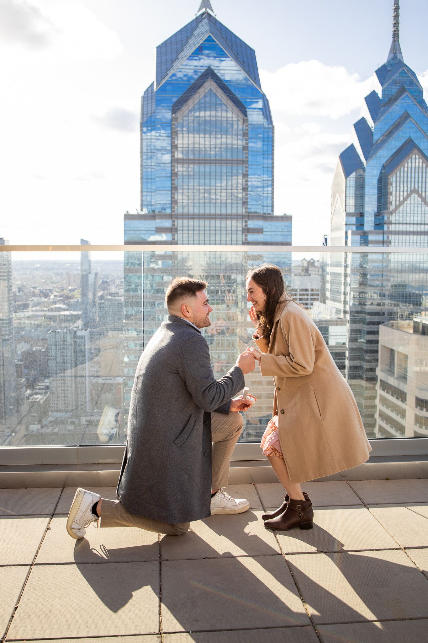 A couple sits together on a rooftop terrace overlooking Philadelphia's iconic blue glass skyscrapers on a sunny day.