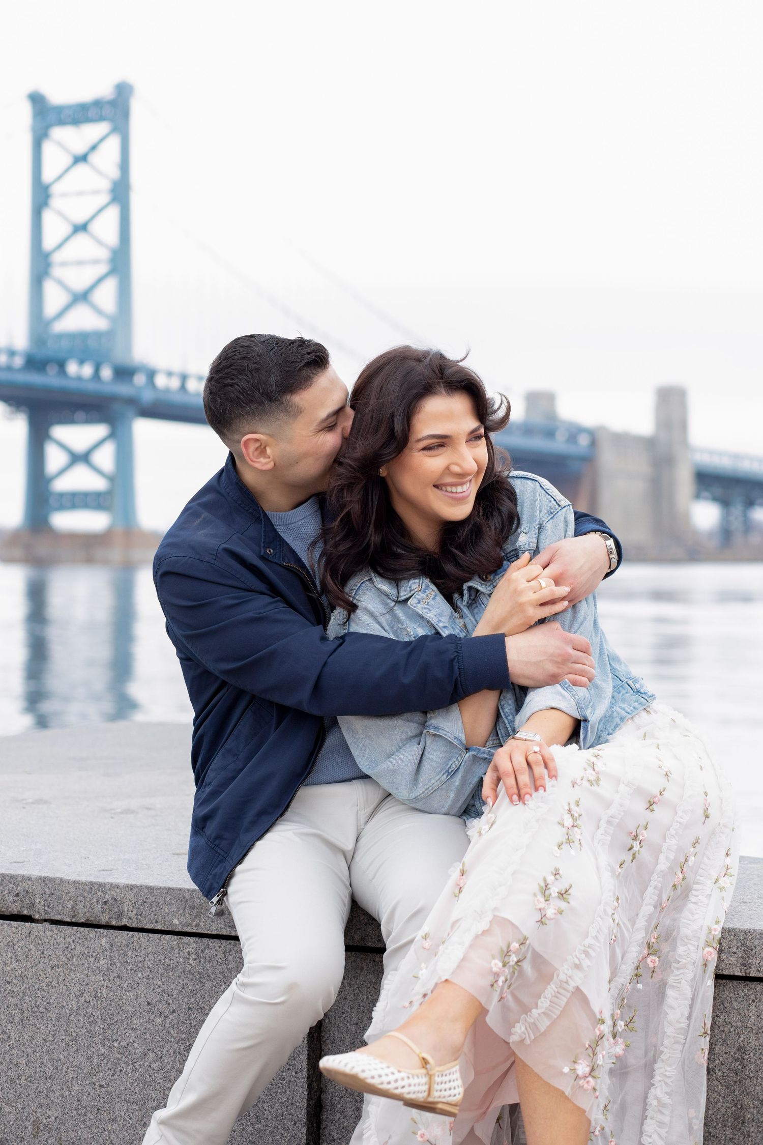 A couple shares a tender moment while sitting by the Ben Franklin Bridge in Philadelphia on an overcast day.