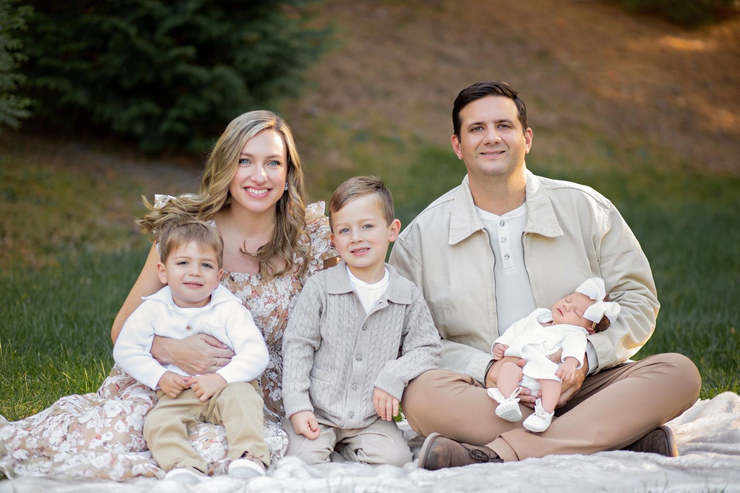 Family of five sitting together outdoors on a blanket in a grassy area with evergreen trees in background.