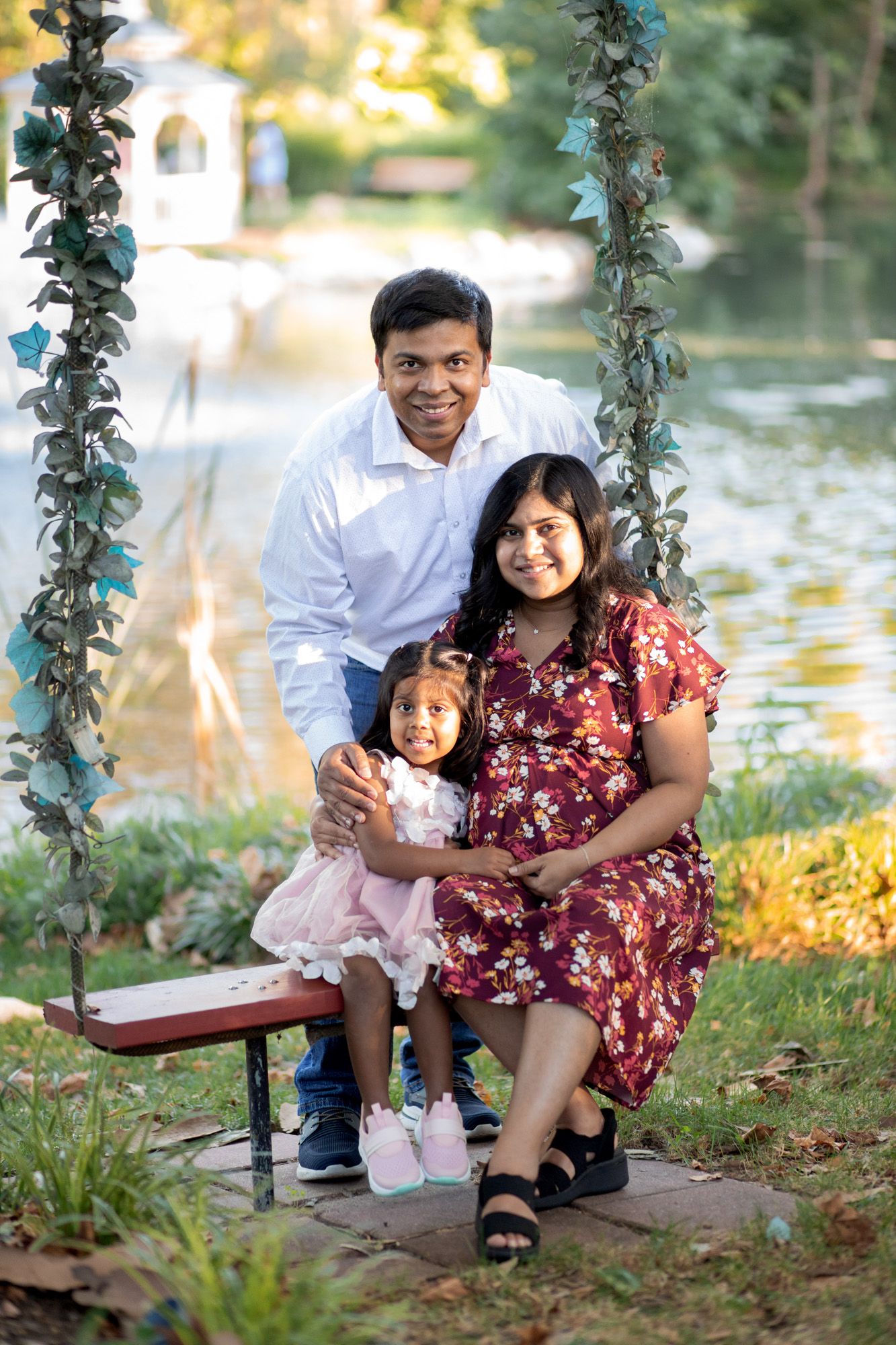 Happy family maternity photo with pregnant mother, father, and toddler daughter on garden swing.