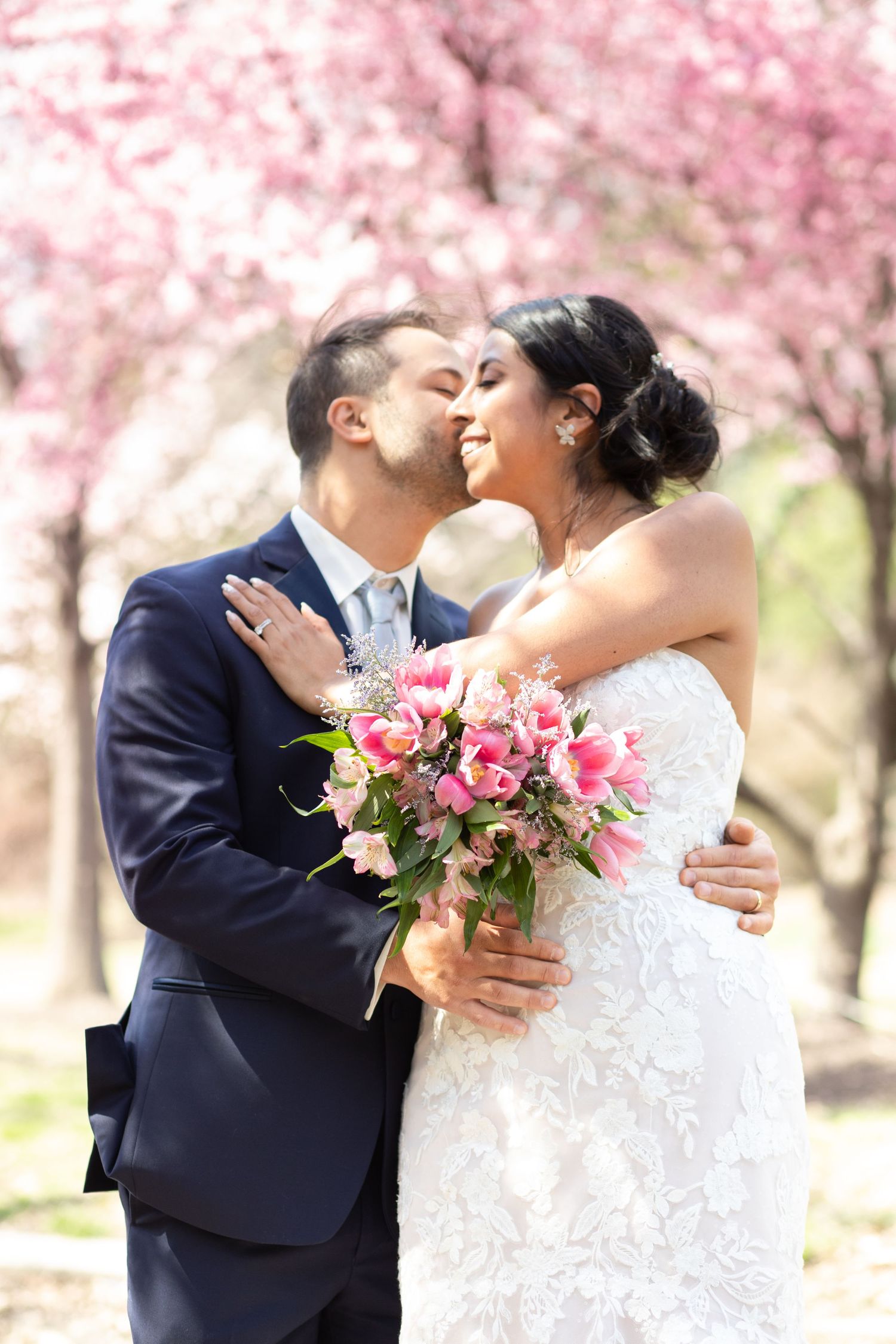 A joyful couple shares an intimate moment under blooming cherry blossoms while holding a pink floral bouquet.