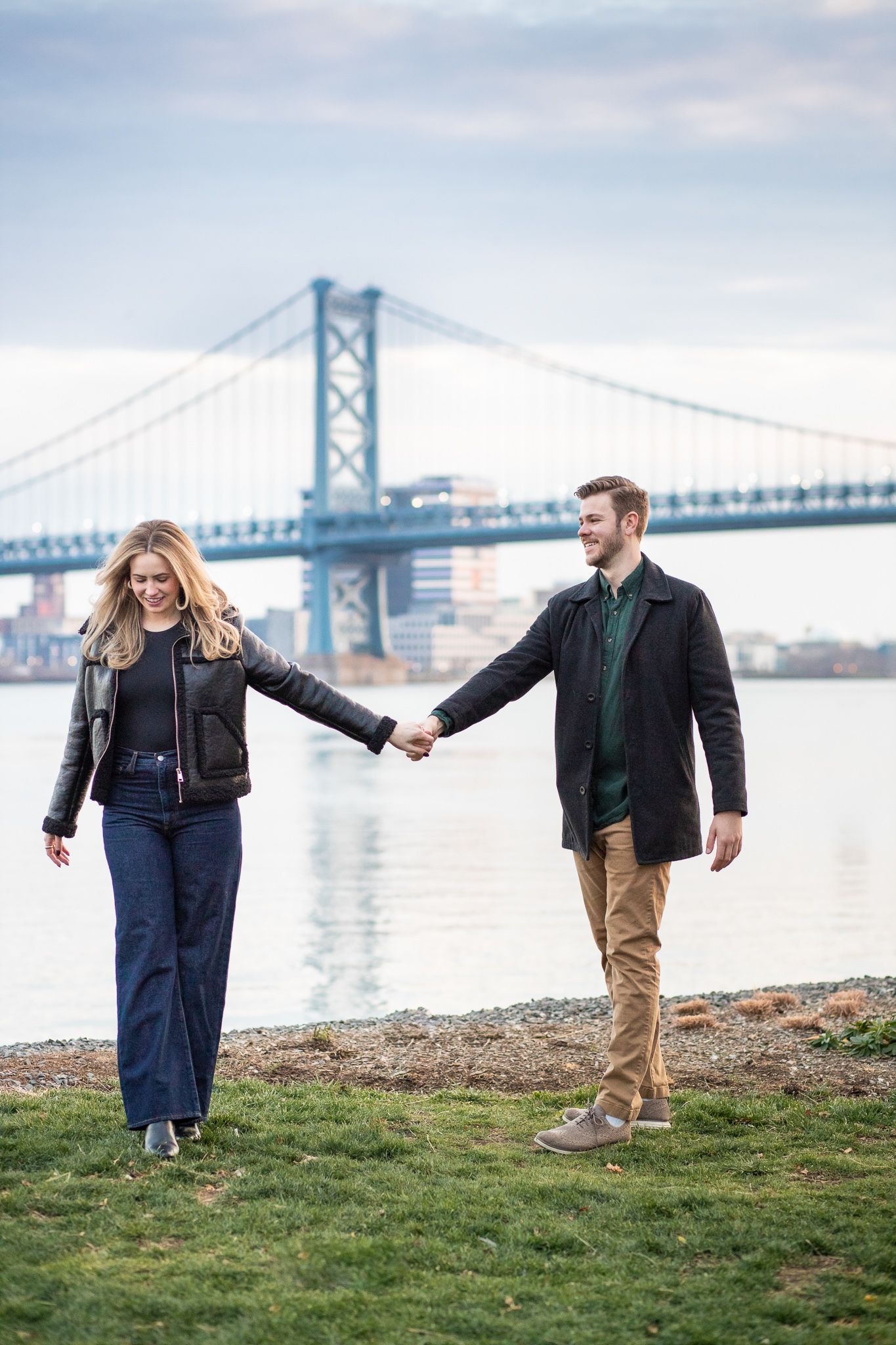 Two people walk and embrace along a grassy riverbank with the Benjamin Franklin Bridge in the background.