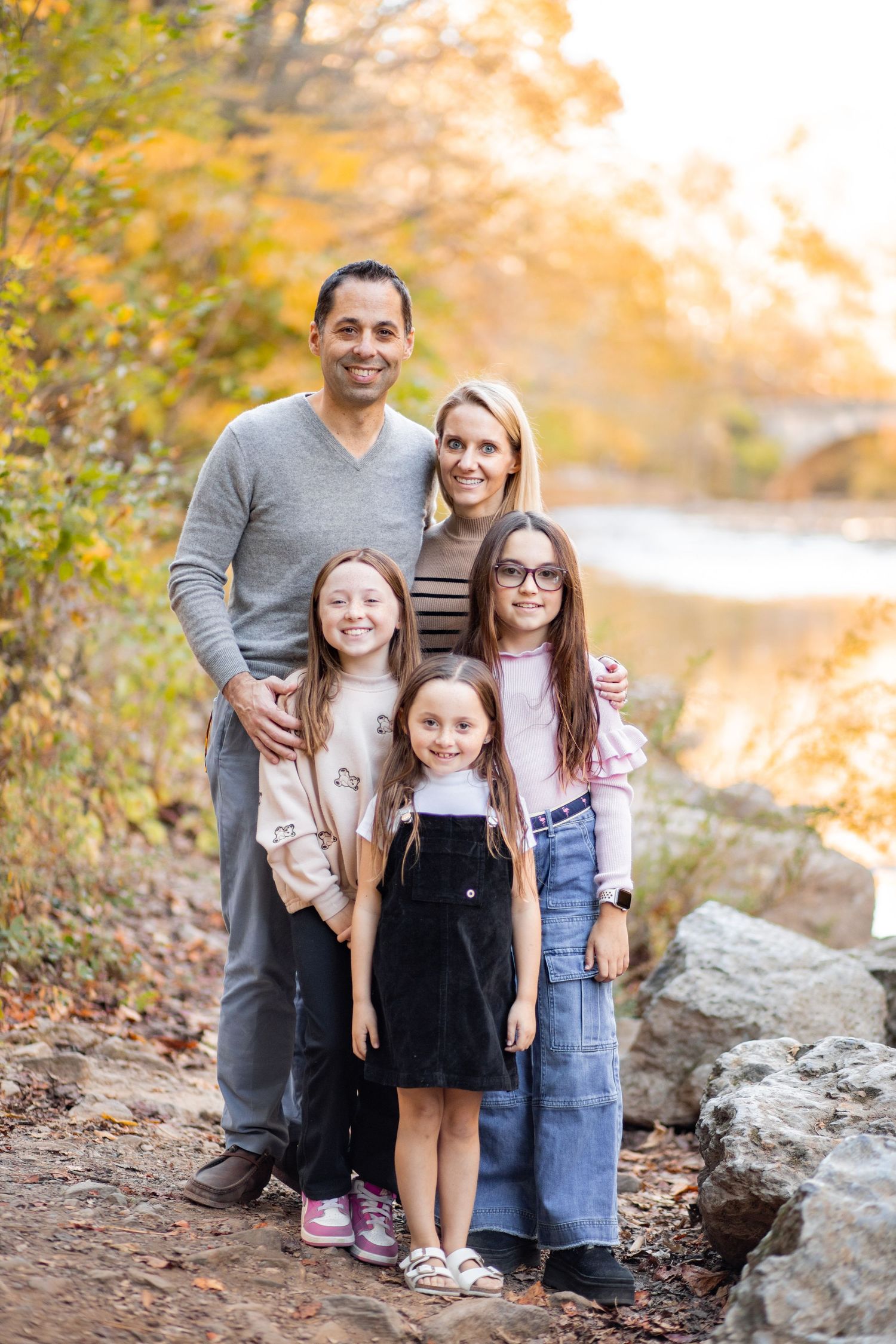 A family poses together on a rocky path during autumn with golden trees and a lake in the background.