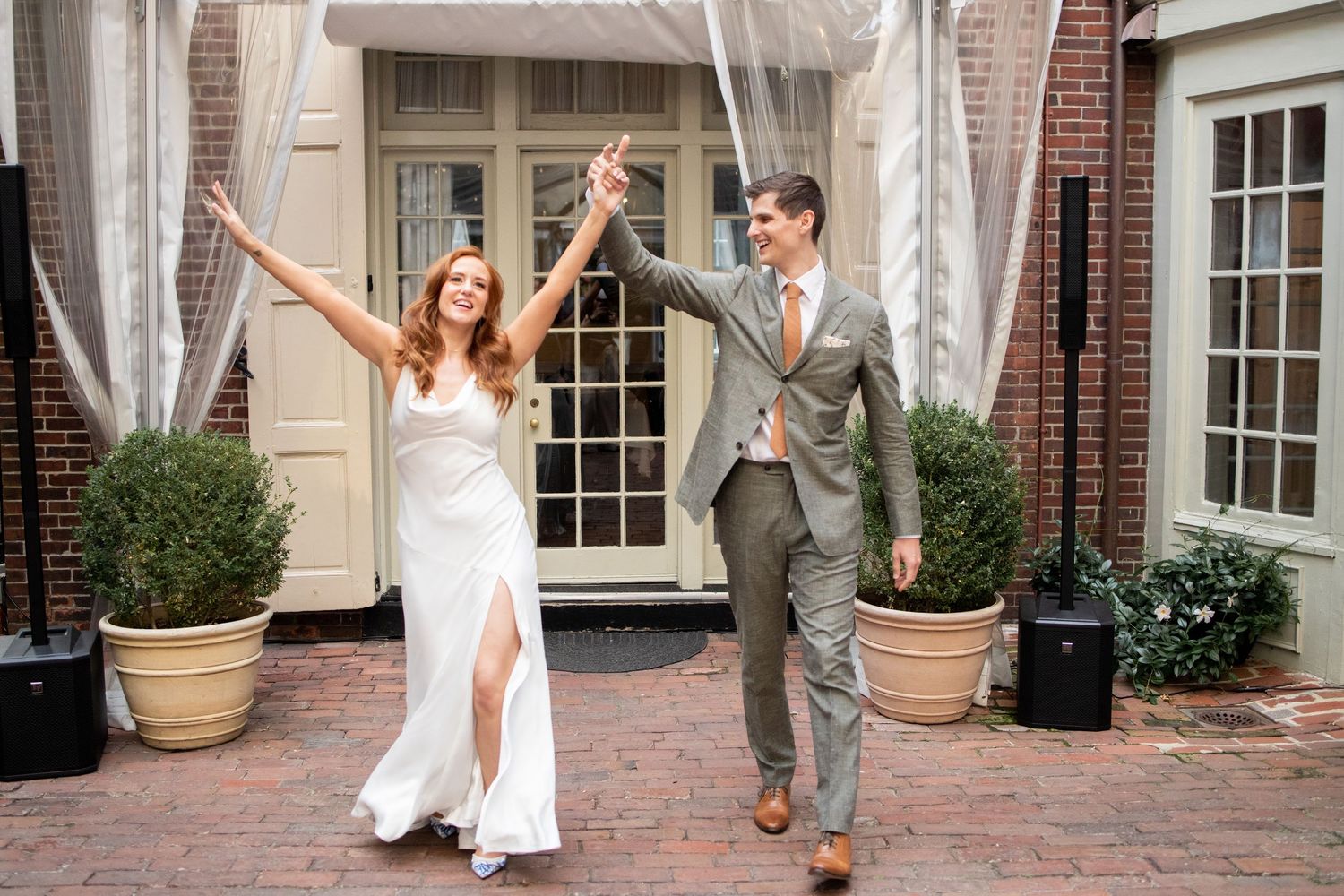 A bride and groom hold hands and celebrate joyfully while walking on a brick courtyard outside an elegant building entrance.