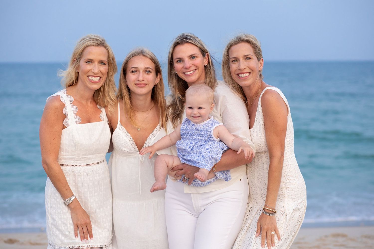 A group wearing white outfits poses together on a sunny beach with turquoise ocean waves in the background.