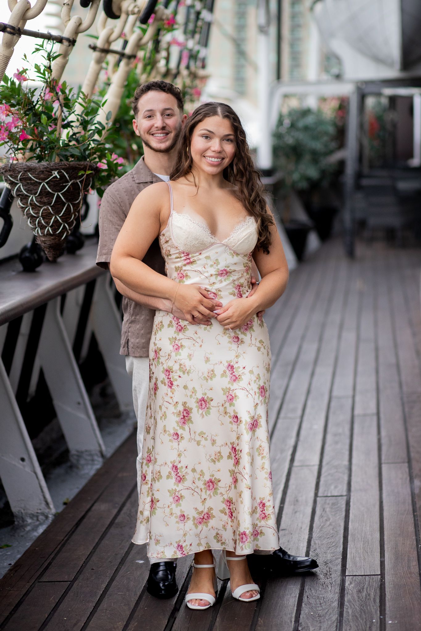 A romantic couple embraces on a wooden deck surrounded by greenery and outdoor decor.