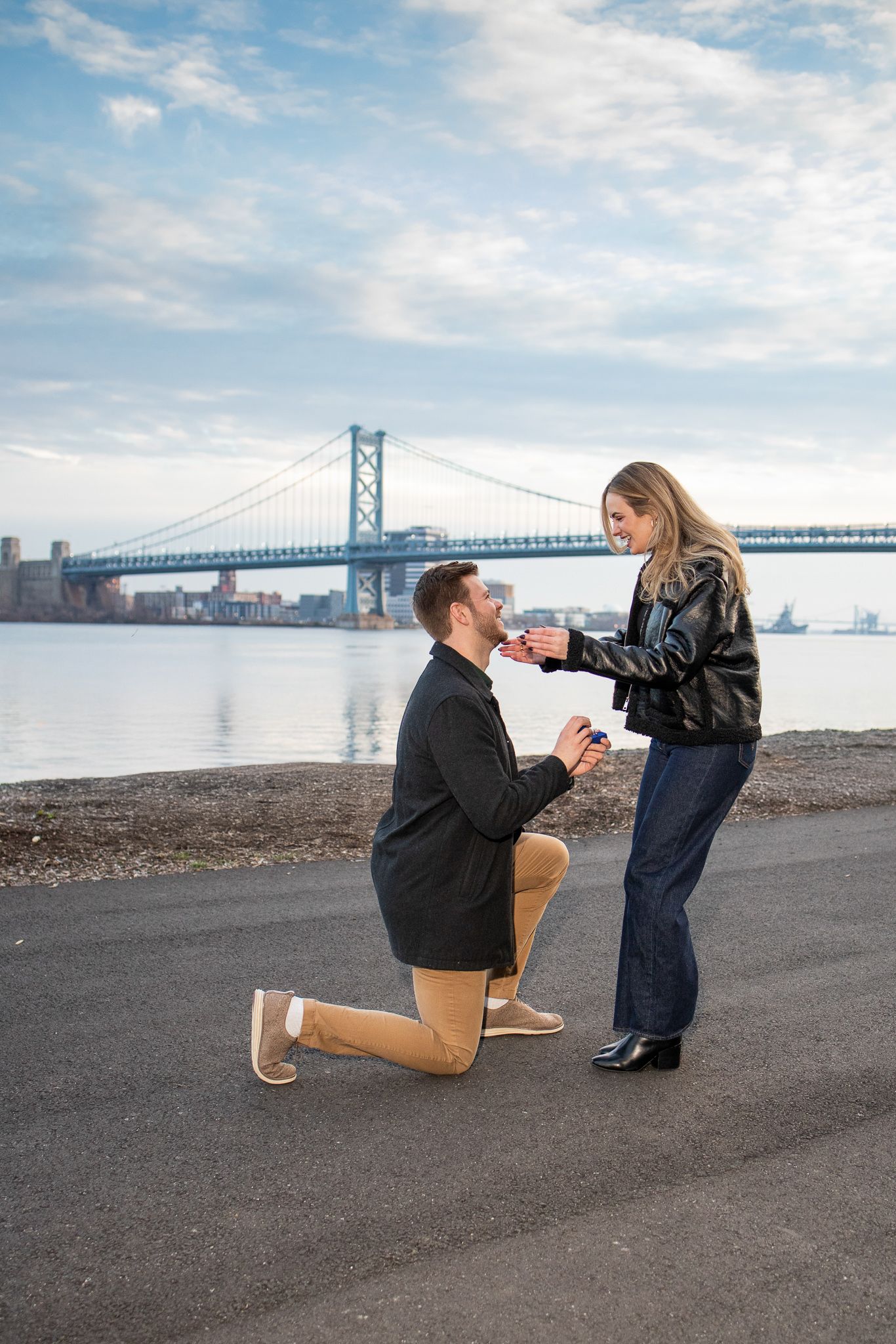 A couple shares a romantic proposal moment by the waterfront with the Benjamin Franklin Bridge in Philadelphia visible.
