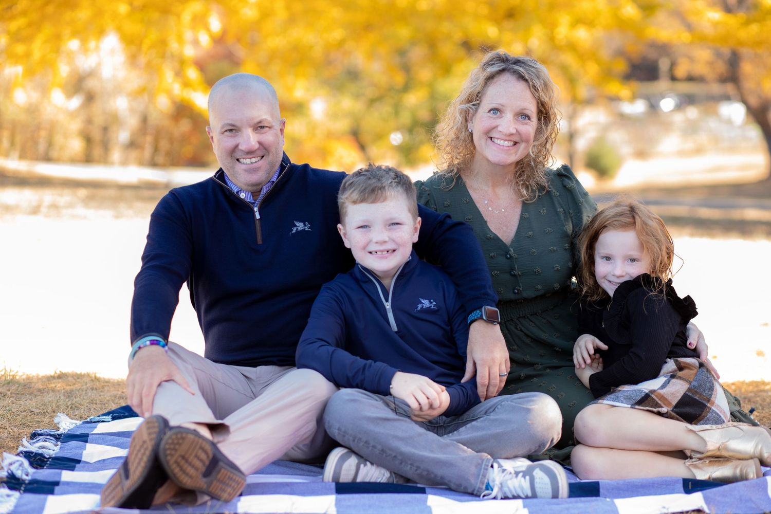 A family poses together on a wooded path during a sunny autumn day with golden foliage in the background.
