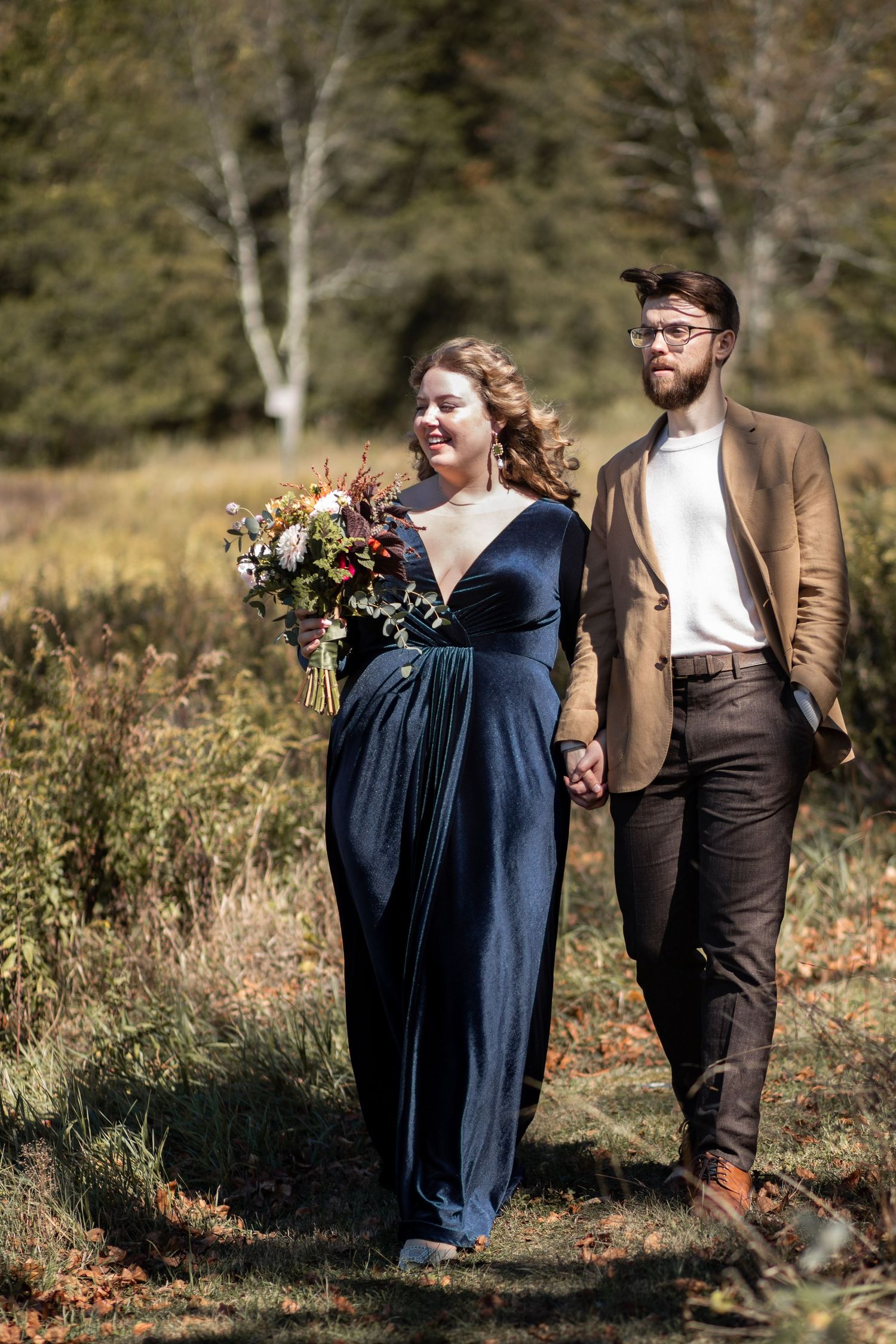 A couple stands in a forest meadow, with the woman wearing a navy dress and holding a bouquet.