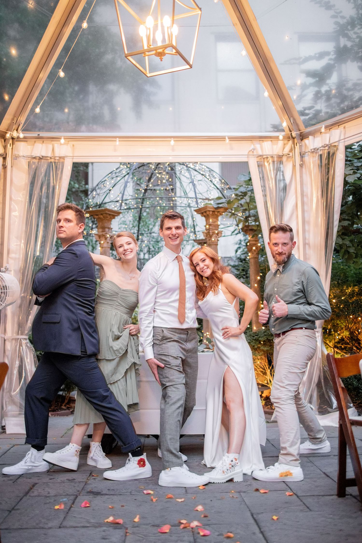 Wedding party poses playfully under an elegant outdoor gazebo with string lights and white drapery.