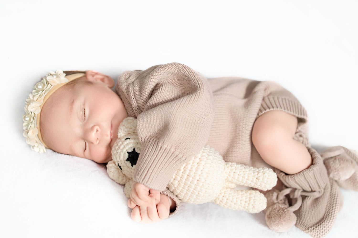 A peaceful sleeping baby wearing a beige knit outfit and lace headband while holding a small stuffed toy.