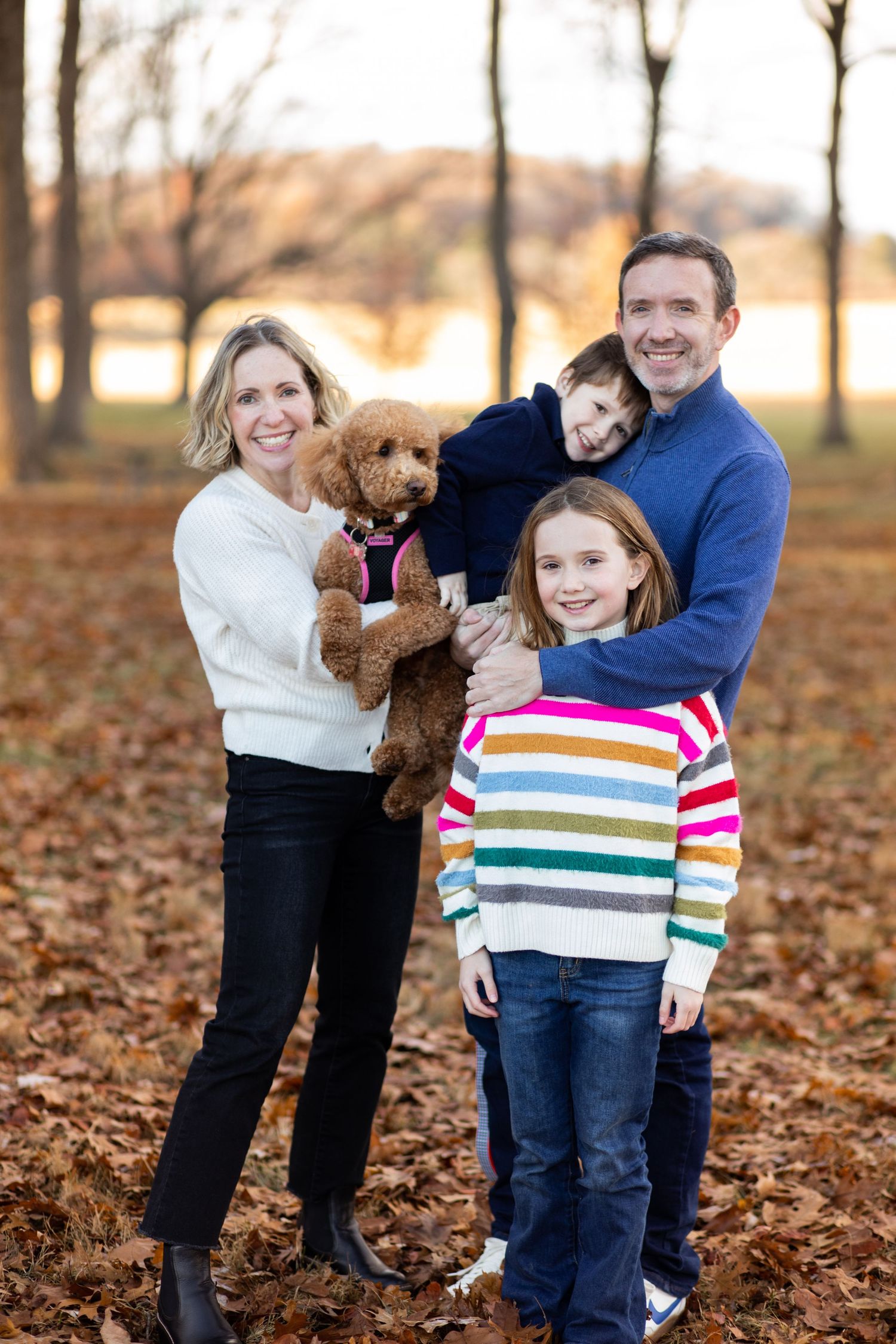 A group portrait on fallen autumn leaves in a park setting with a small brown dog and colorful striped sweater.