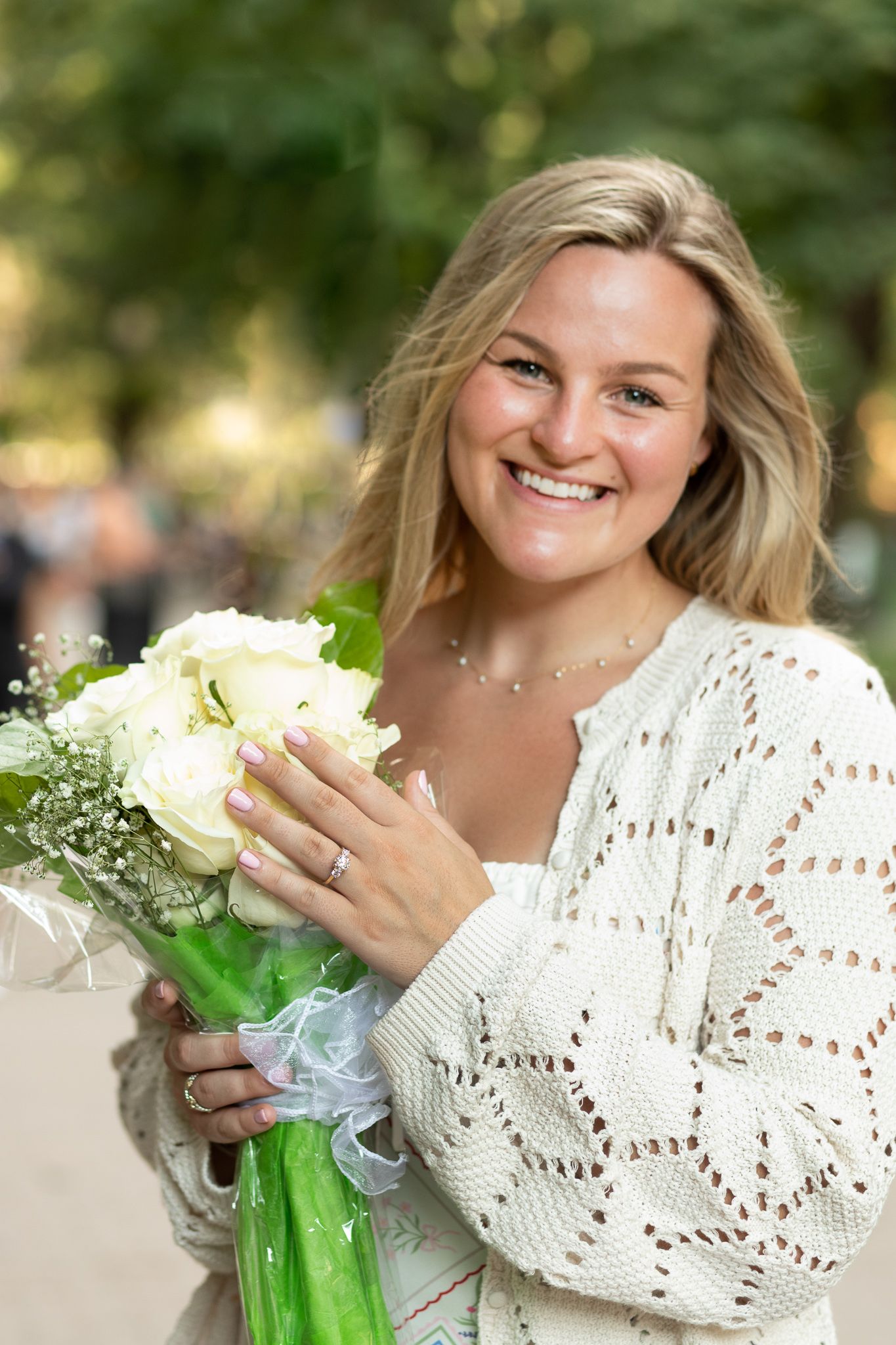 A person in a white crochet sweater poses with white flowers in an outdoor setting during a sunny day.