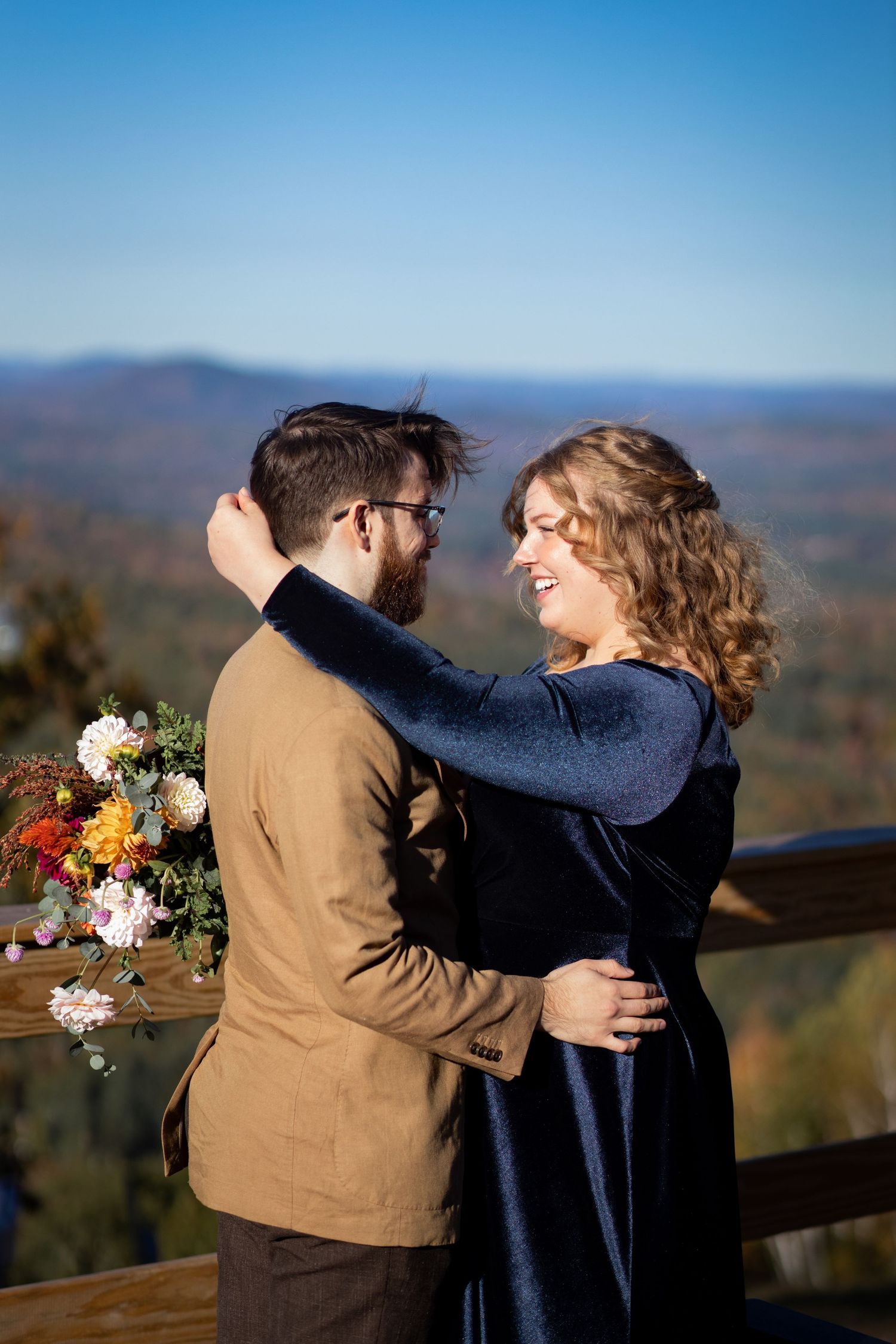 A couple embraces on a mountain overlook with fall foliage and rolling hills in the background during golden hour.