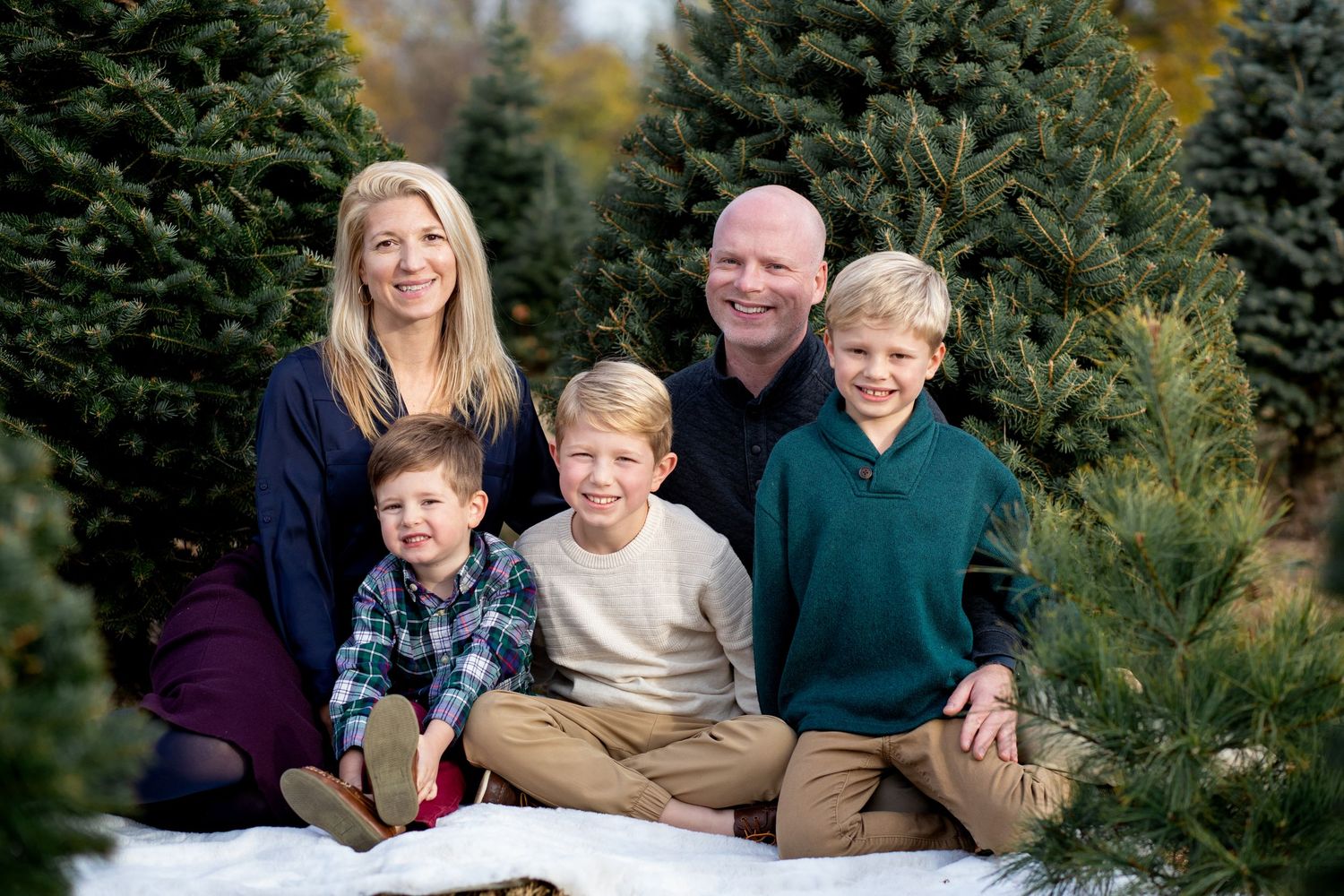 Family of five sitting together and smiling at a Christmas tree farm on a blanket during autumn season.