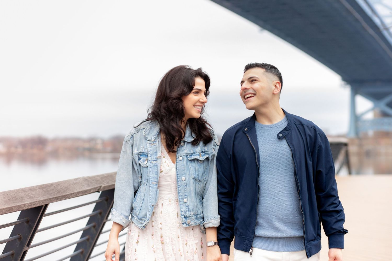 Two people laugh and smile together while walking along an urban waterfront with a bridge in the background.