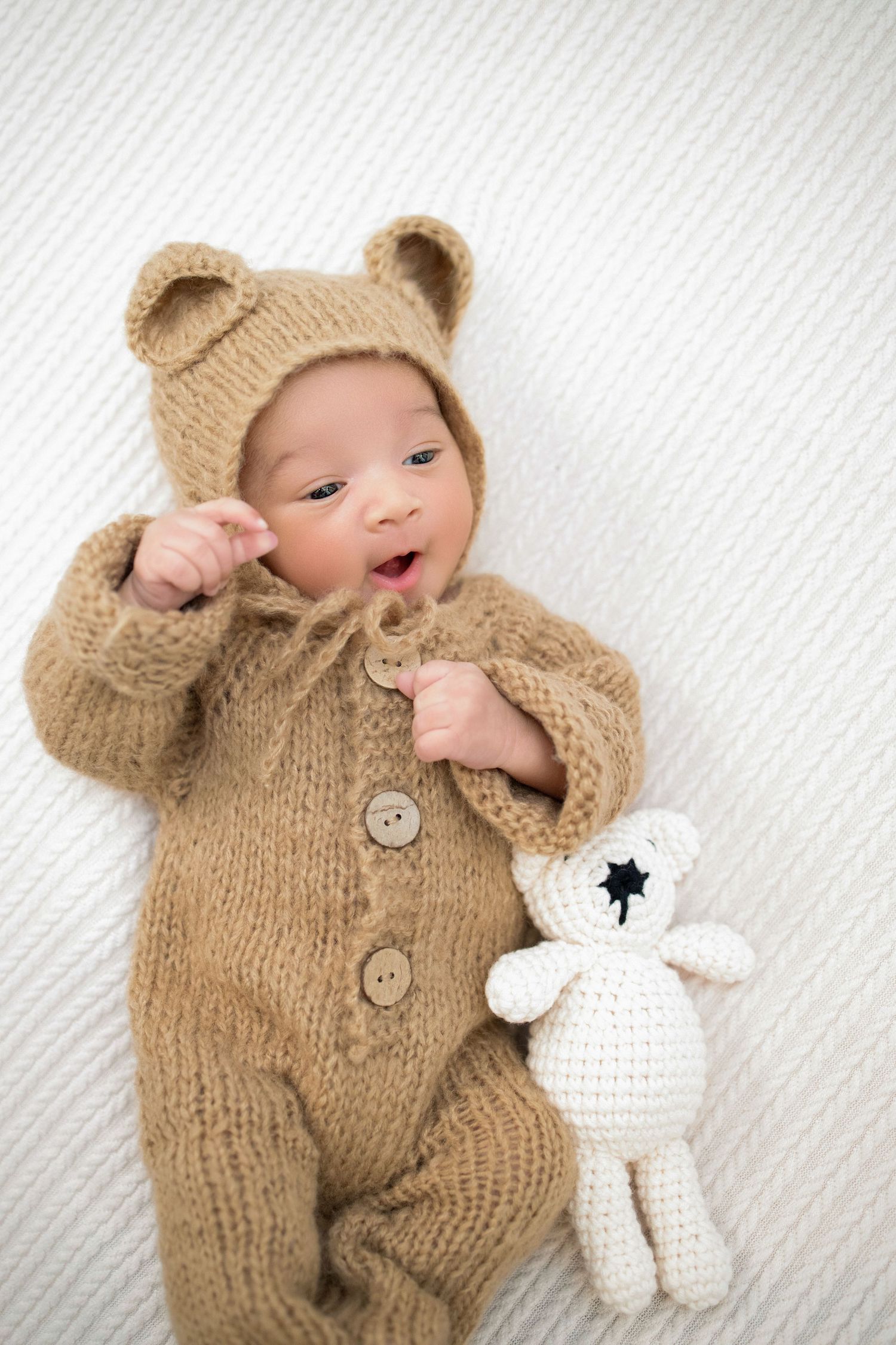 A small baby wearing a knitted tan bear outfit lies on white fabric next to a crocheted white stuffed toy.