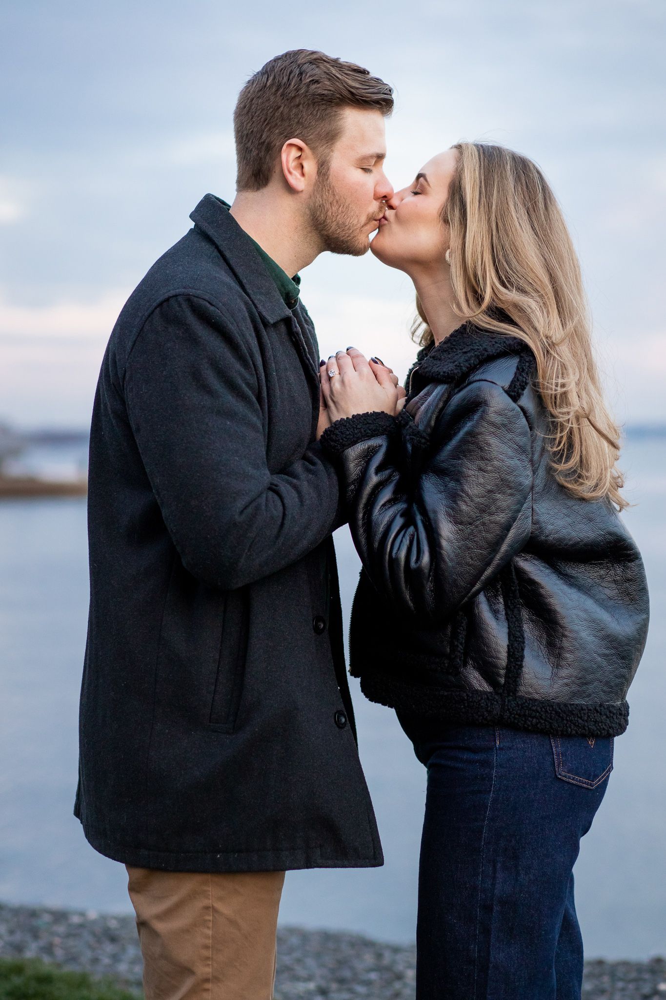 A couple shares romantic kisses against a waterfront backdrop during sunset, creating intimate engagement photos.