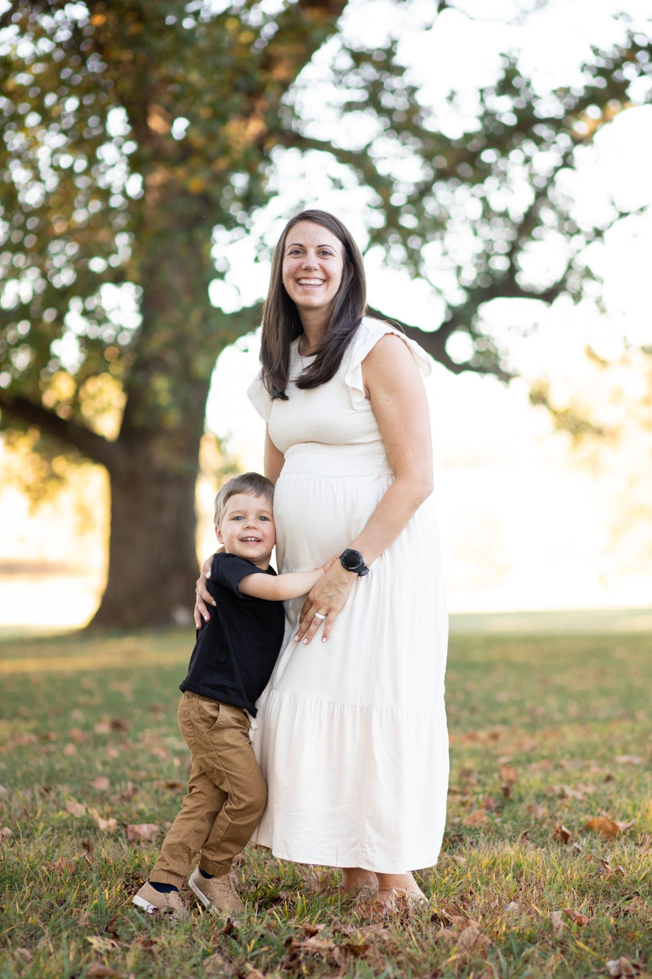 Pregnant woman in white dress smiling outdoors while young son hugs her baby bump.