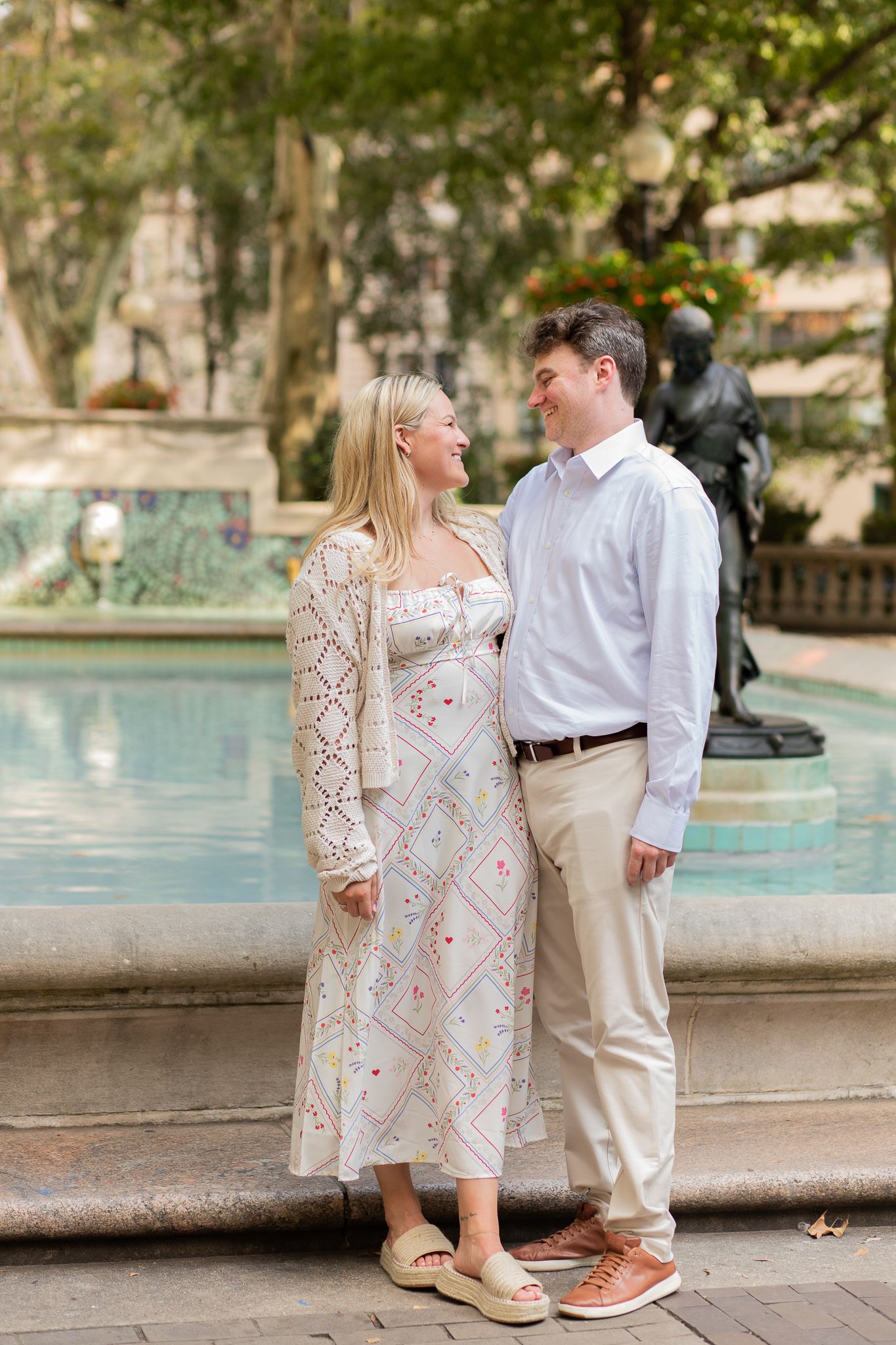 A couple stands together by a fountain wearing coordinated white and beige outfits for engagement photos.