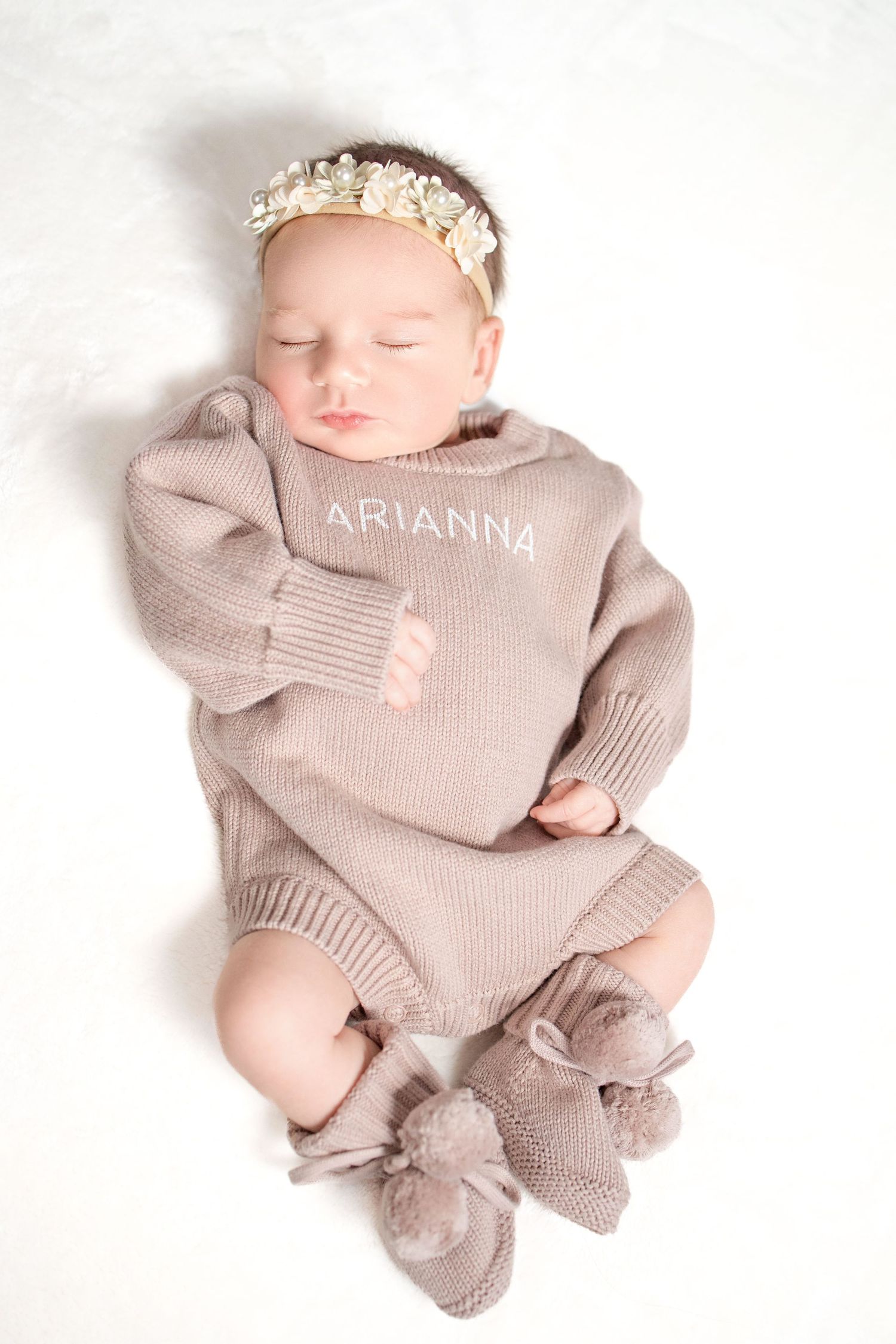 Sleeping baby wearing knit beige sweater with name Arianna and matching booties on white background.