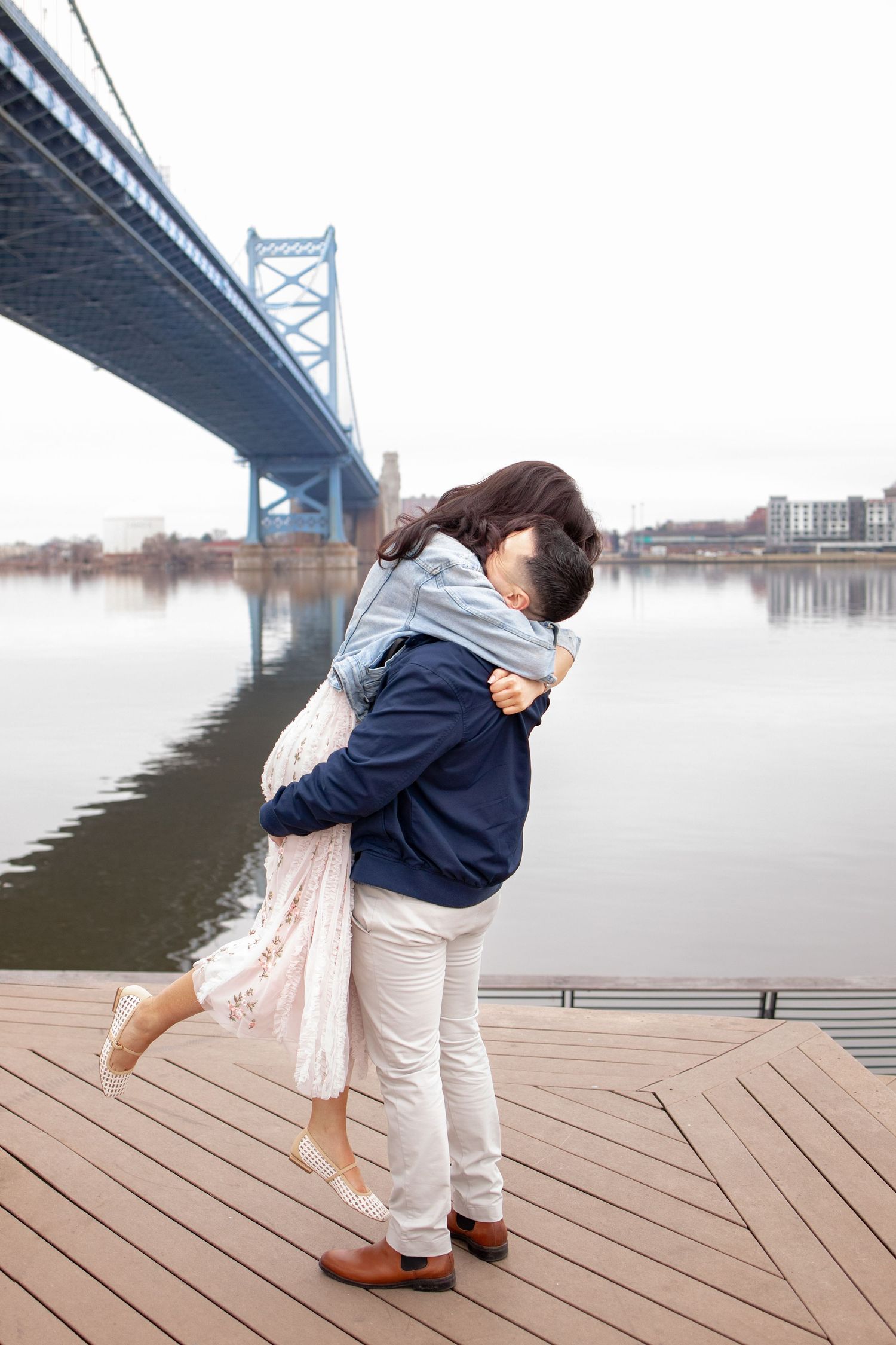 A couple embraces in a romantic moment on a wooden deck beneath the Benjamin Franklin Bridge in Philadelphia.
