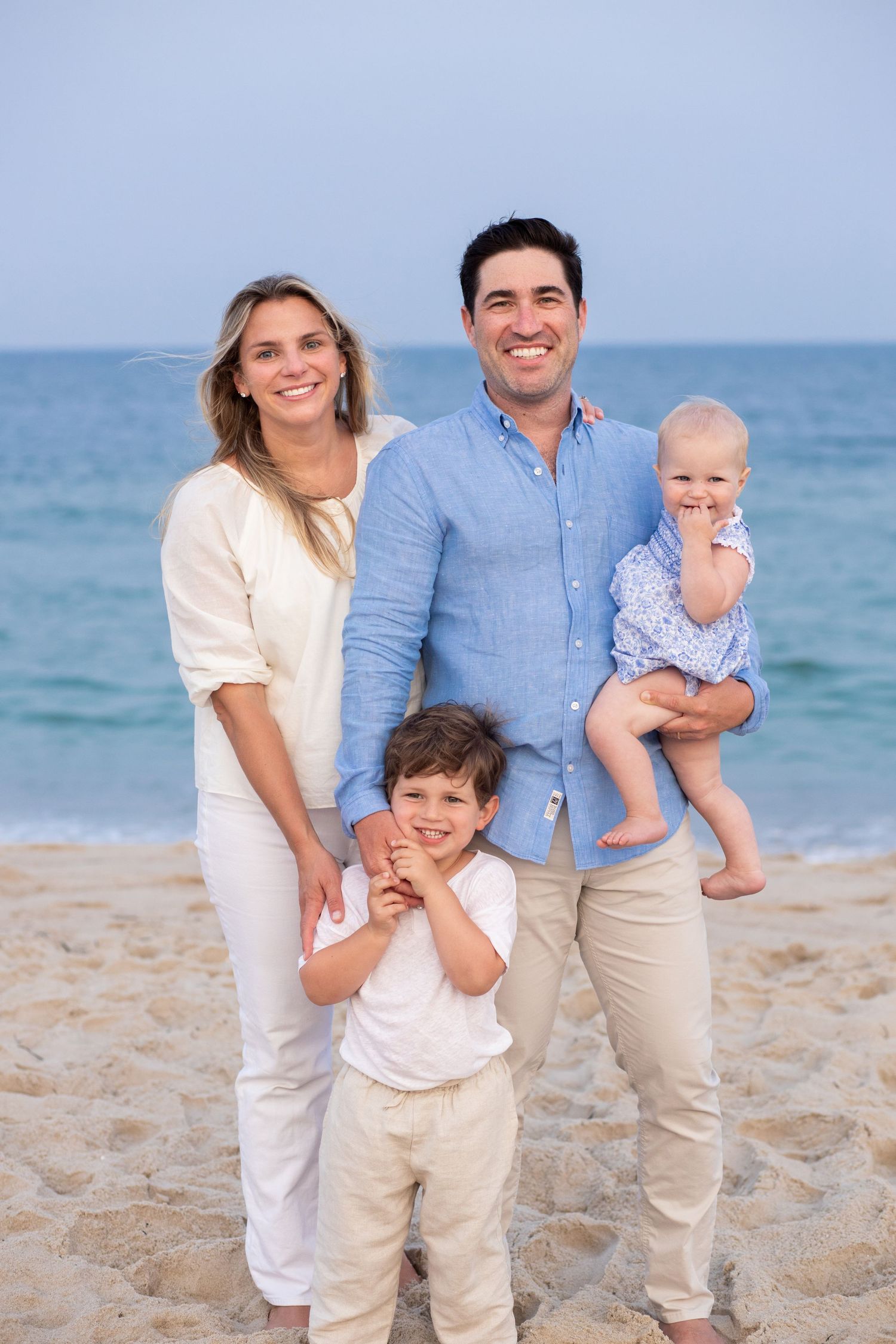 Family wearing coordinated white and blue outfits poses together on sandy beach with ocean waves in background.