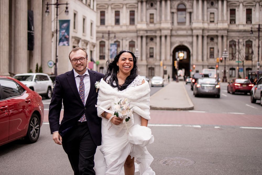 A couple in wedding attire share a joyful moment walking together near Philadelphia City Hall on their wedding day.